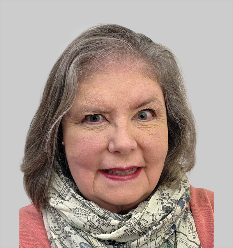 Close-up of an older woman with shoulder-length gray hair, blue eyes, wearing a patterned scarf and a pink top, smiling against a black background.