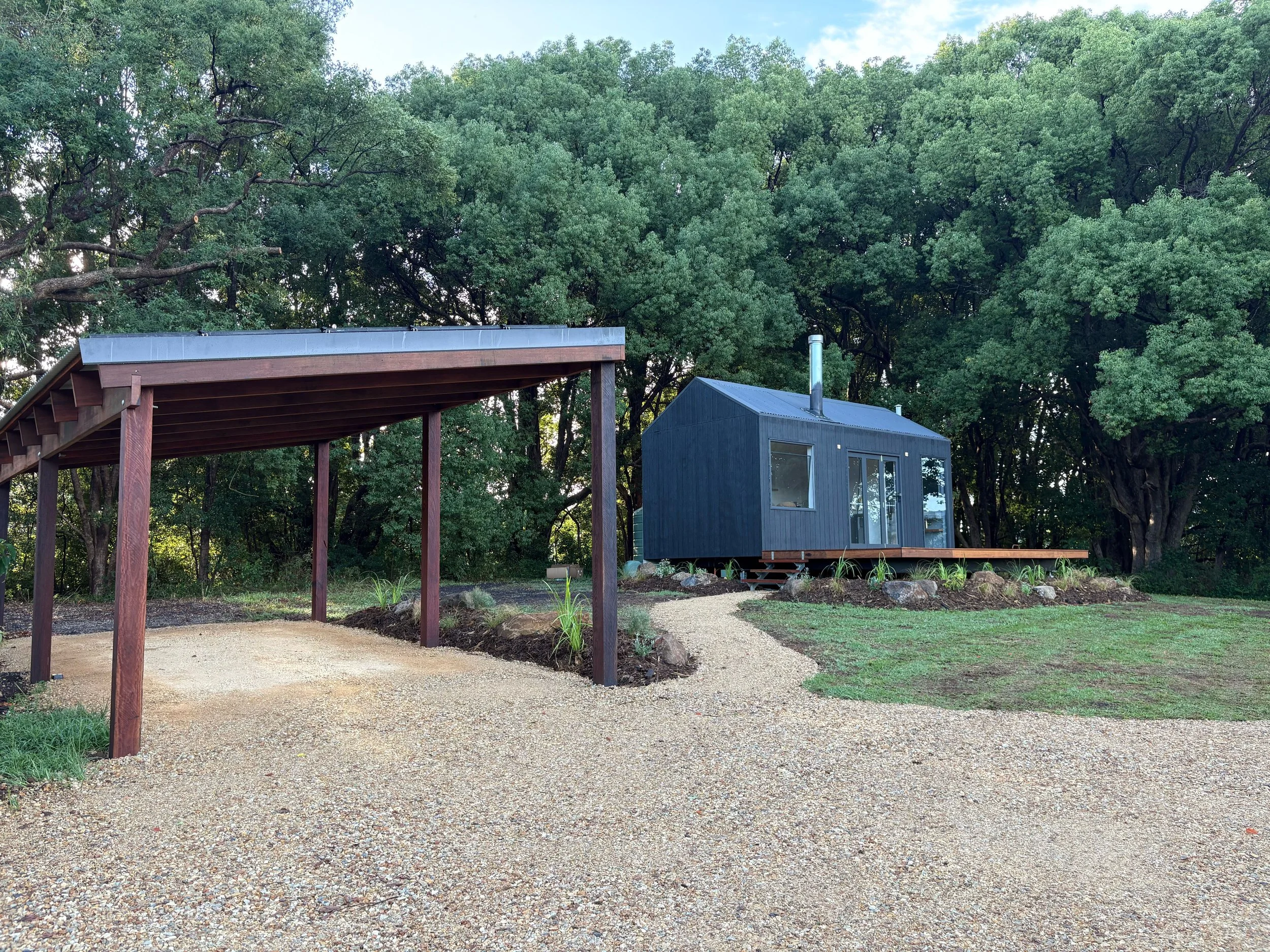 A small, black tiny house on a wooden platform surrounded by trees, with a gravel path leading to it and a carport on the left.