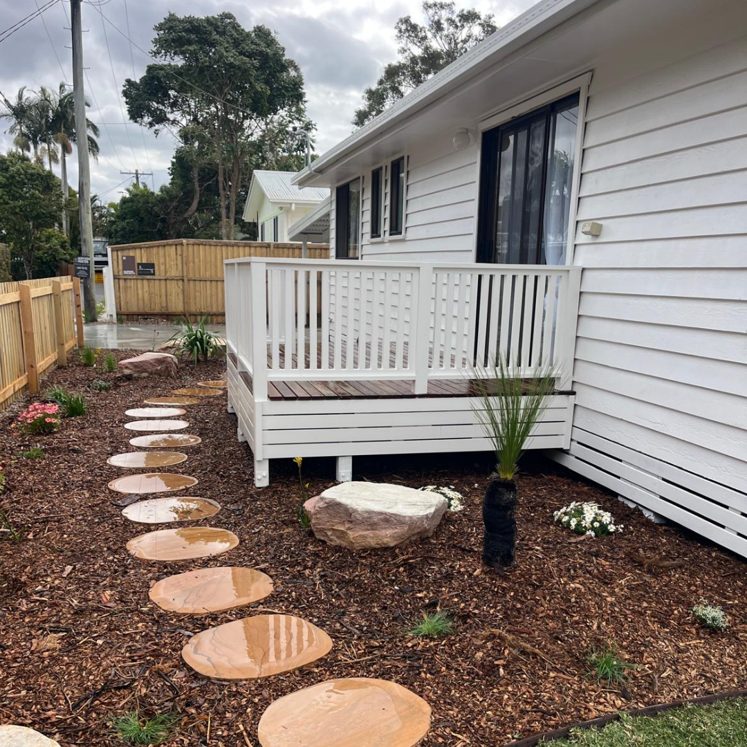 A white house with a small wooden porch and a mulched yard with stepping stones, plants, and rocks, in a residential neighborhood with trees and cloudy sky.