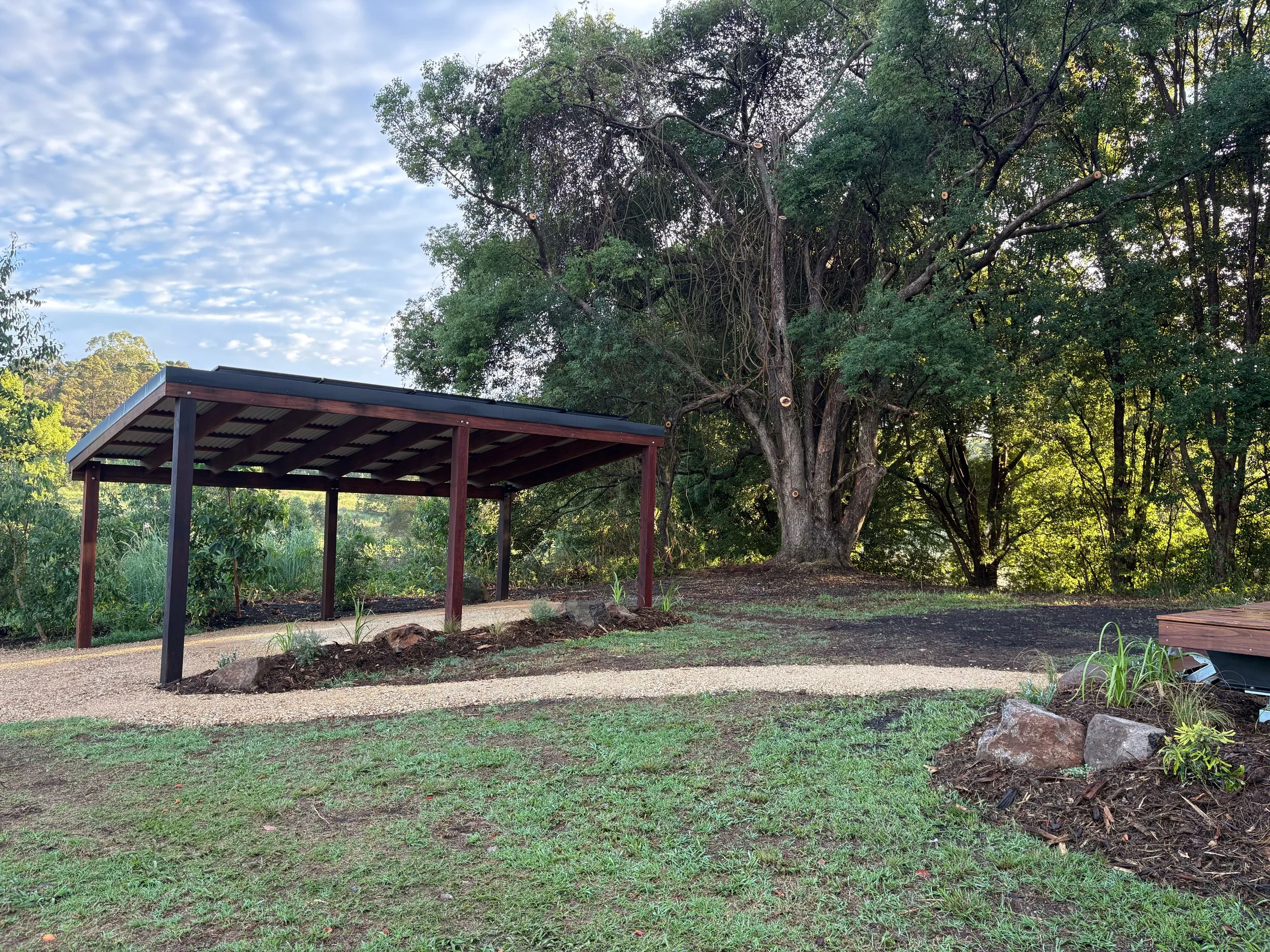 A outdoor scene with a wooden pergola, a gravel pathway, and large trees with green foliage during daytime.