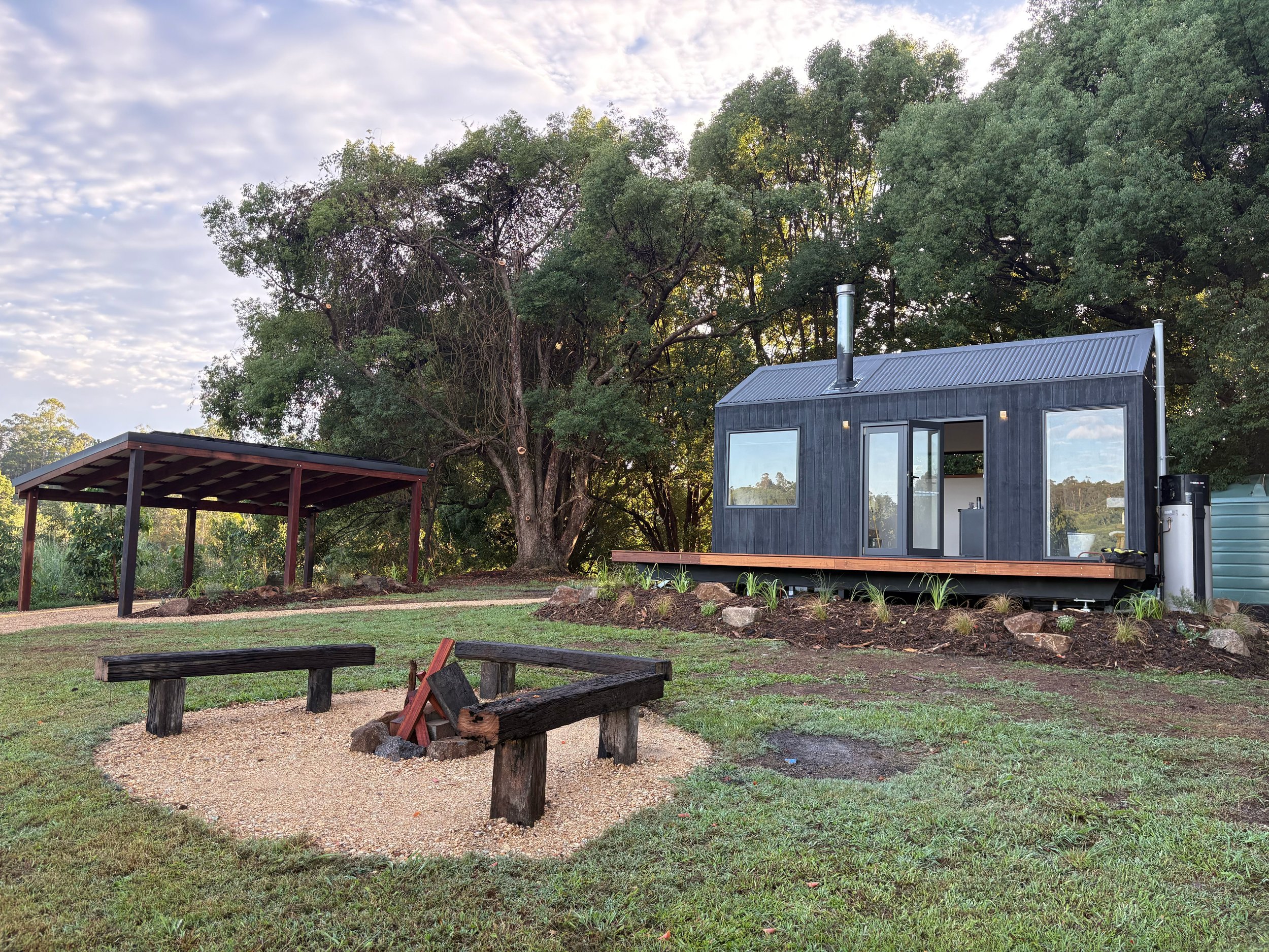 A tiny black cabin with a sloped roof and large windows, situated on a grassy yard with a firepit surrounded by two wooden benches, a large tree, a covered picnic area, and a water tank, under a partly cloudy sky.