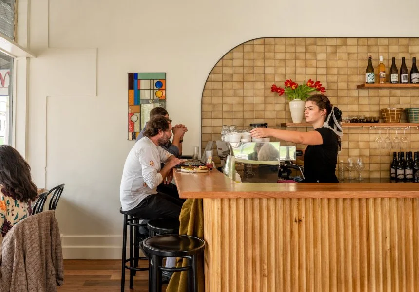 A woman serving coffee behind a wooden counter in a cafe with customers seated at the bar, including a man in a white shirt and a woman in patterned clothing, with a brown tiled wall and framed artwork behind her.