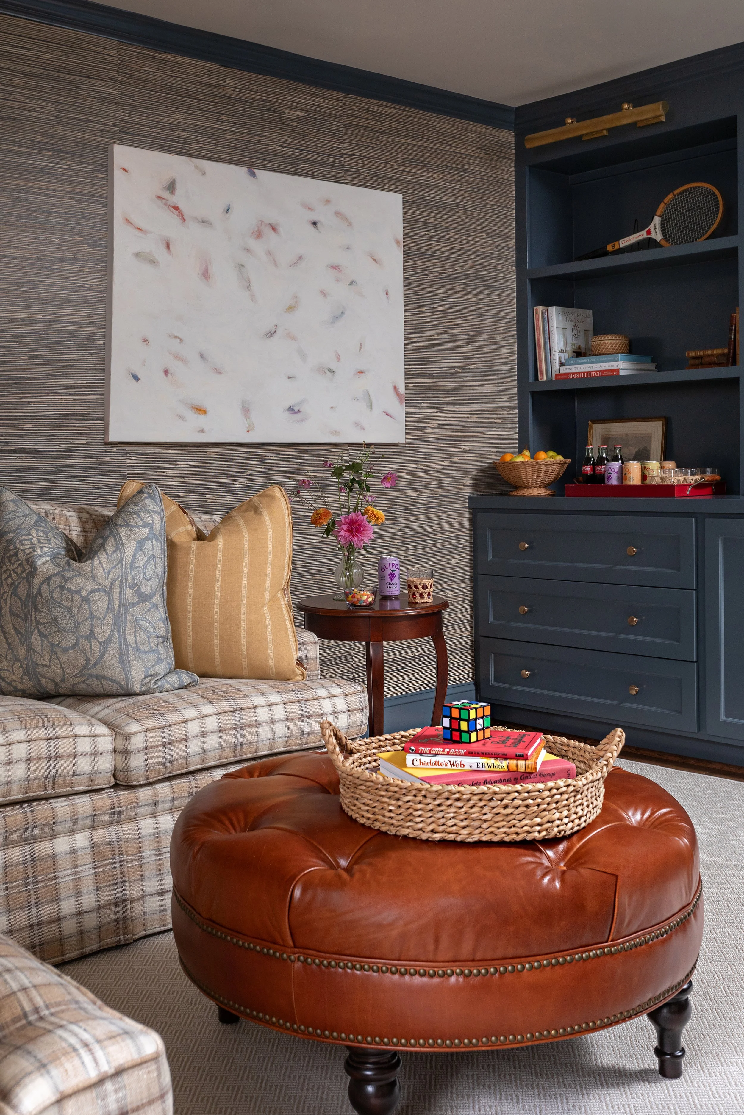 Layered family room with plaid upholstery, cognac leather ottoman, grasscloth walls, and blue built-in cabinetry.