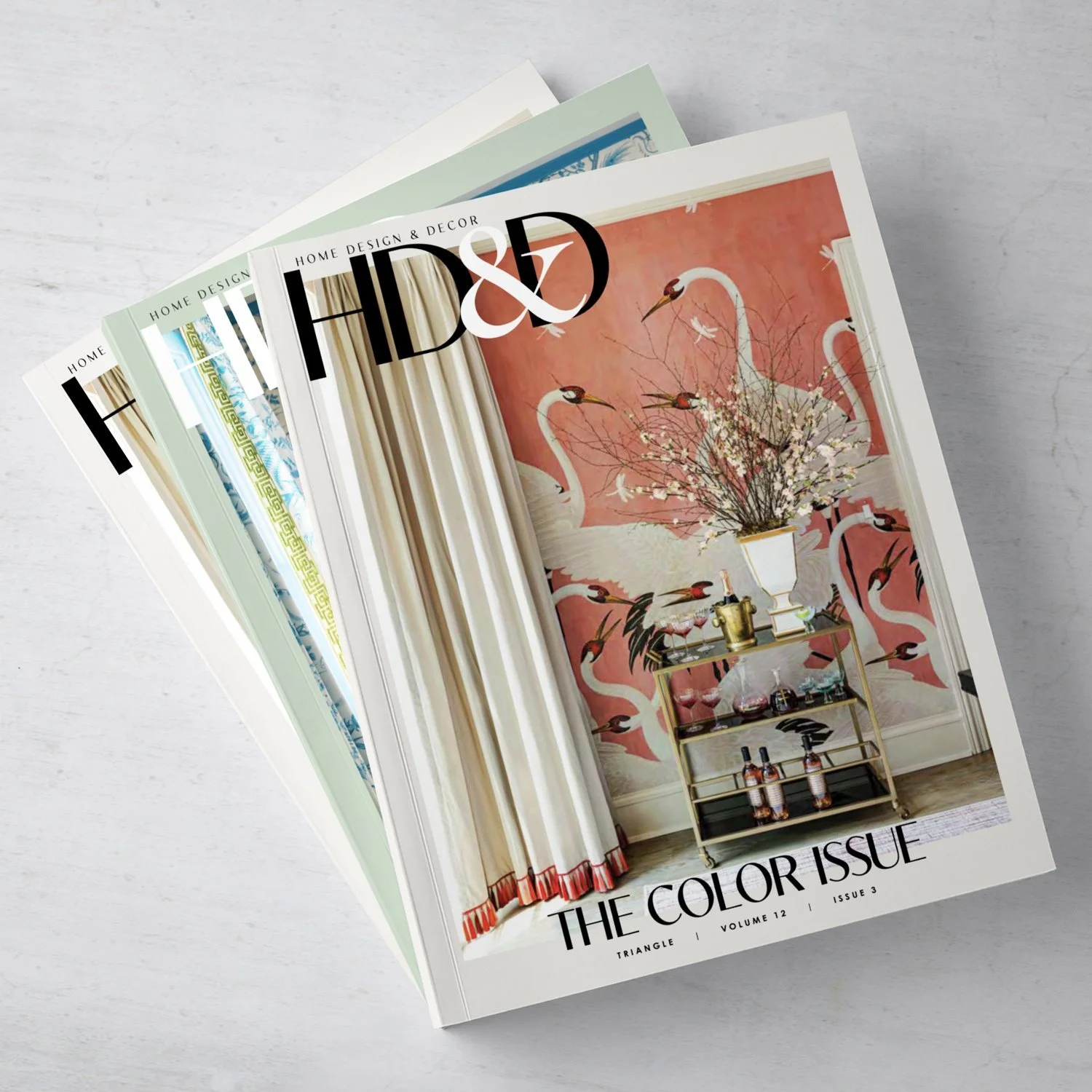 Set of interior design magazines with the top magazine showing a wall with white swan murals and a floral arrangement in a white vase on a gold cart.