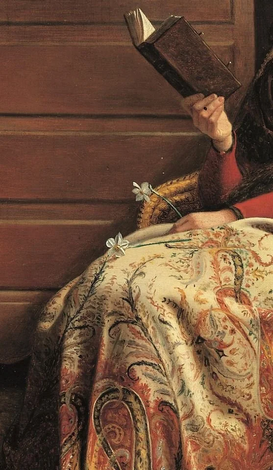 A person sitting on a wicker chair, wearing red and black clothing, holding a book, with their hand resting on a floral-patterned fabric, and white lilies placed on the fabric.