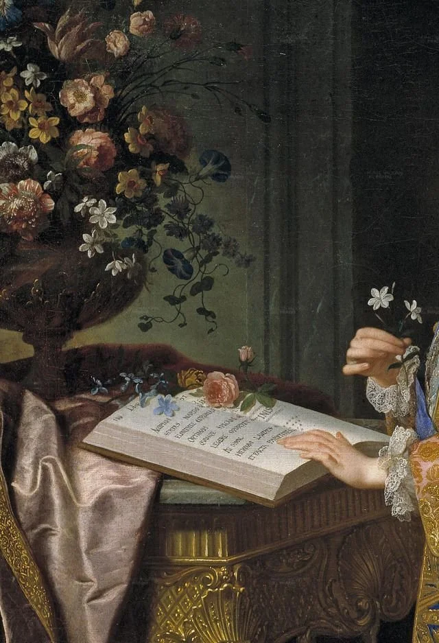 Close-up of a person with lace sleeves holding a flower, reading a large open book on a decorative wooden table, surrounded by a floral arrangement in a vase.
