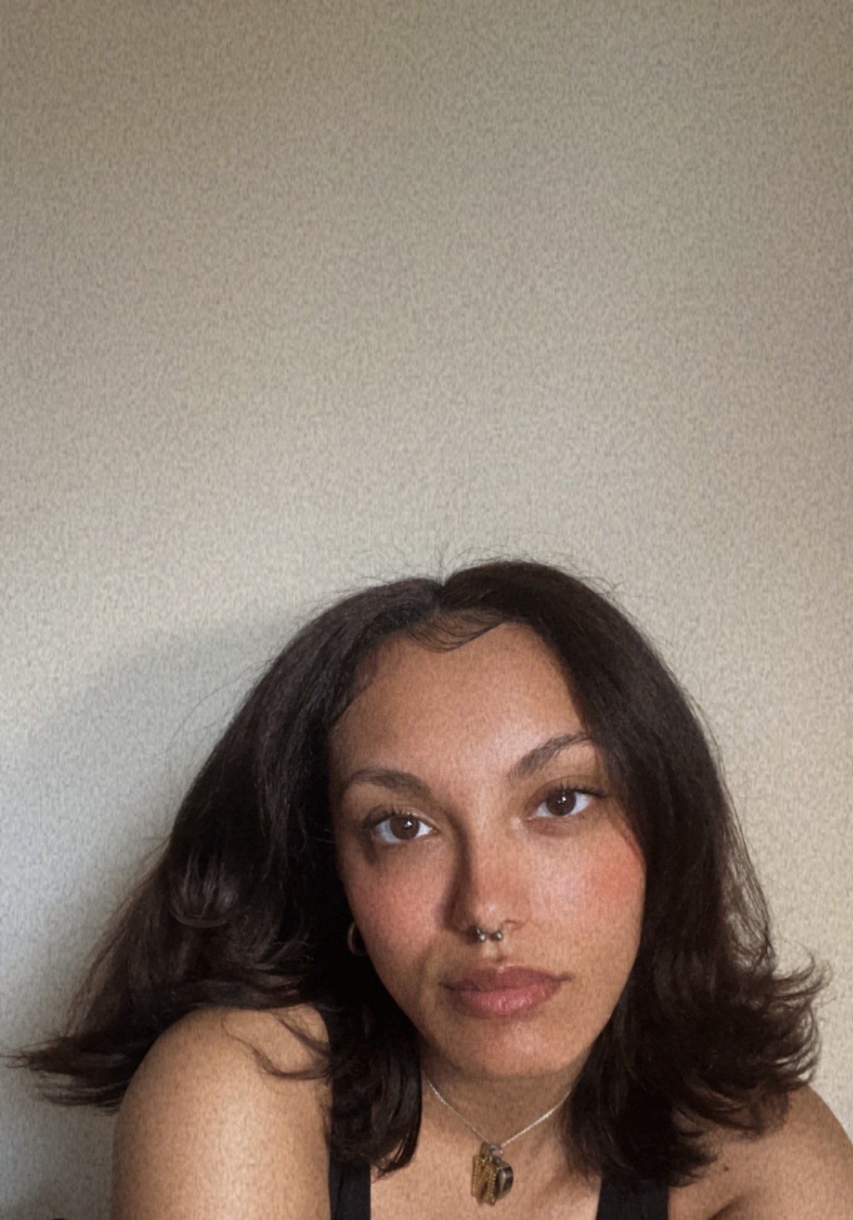 Close-up of a young woman with wavy, shoulder-length dark hair, wearing a septum piercing, a necklace with an ornate pendant, and hoop earrings, against a plain beige wall.