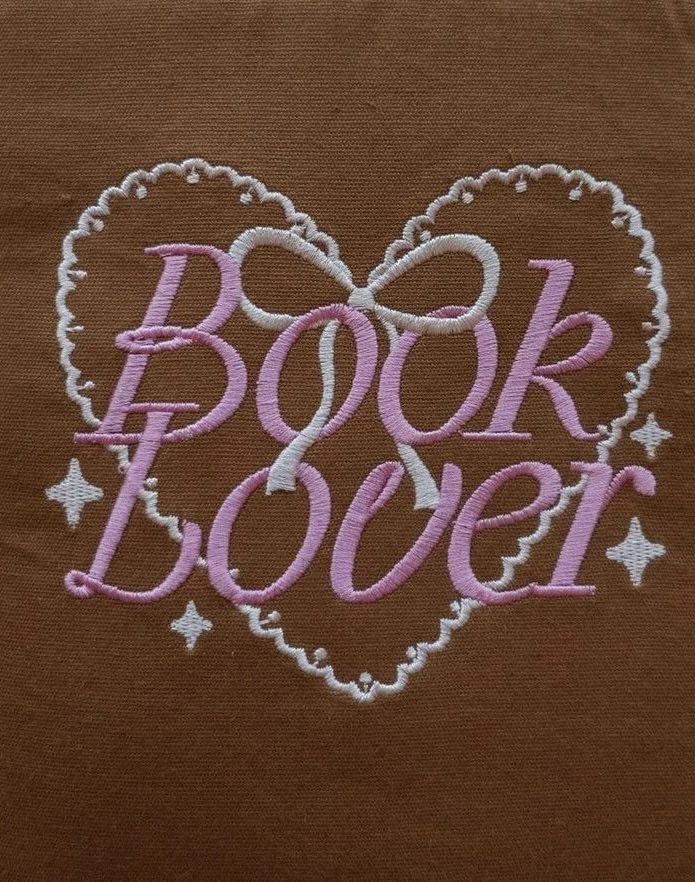 Embroidery design on fabric featuring a heart shape with the words ' BOOK BOUER' in pink and white thread, surrounded by stars.