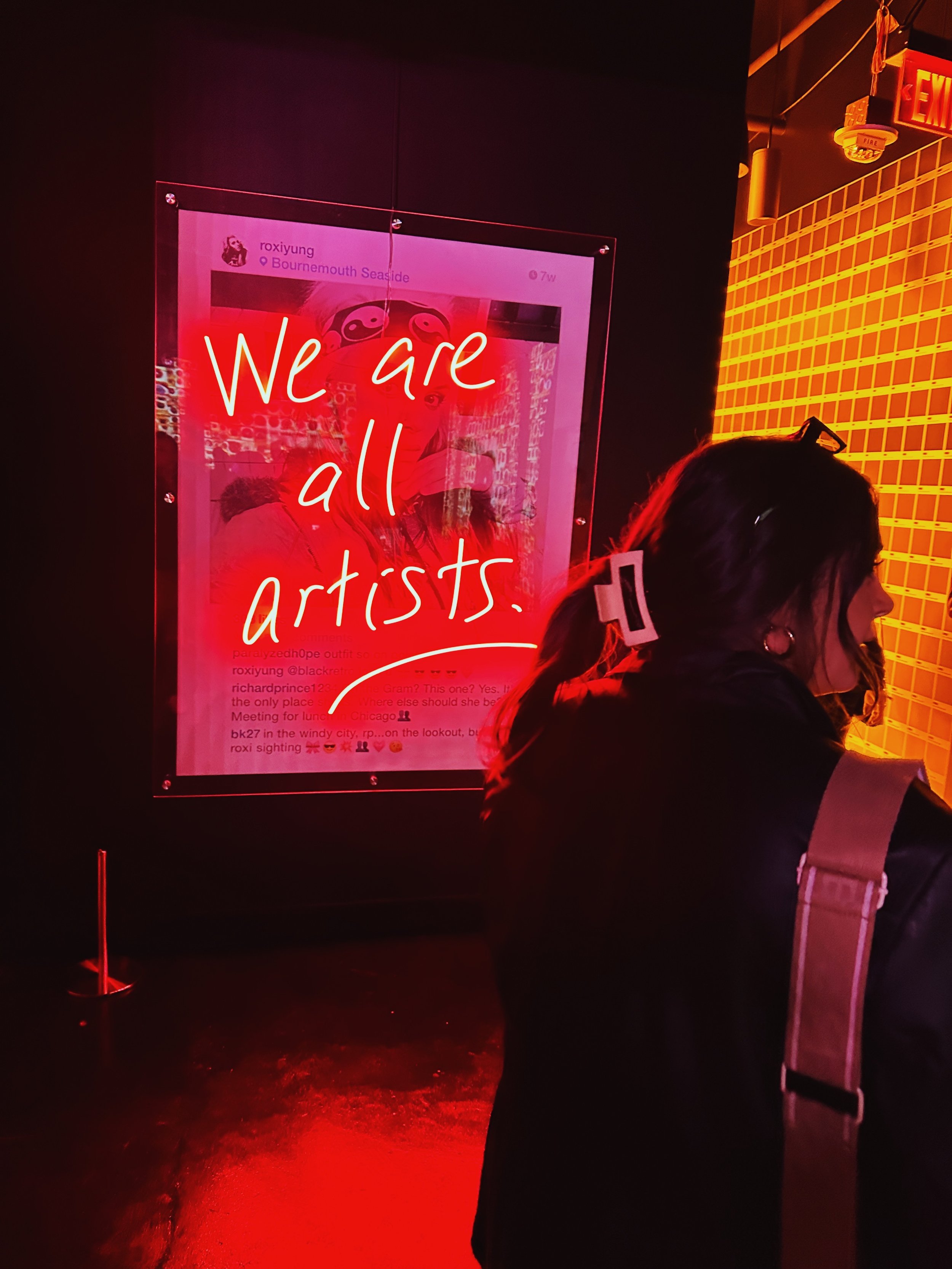A neon sign with red and white text reading 'We are all artists' displayed on a glass panel. Nearby is a person with dark hair and glasses, wearing a beige bag, standing in a dark indoor space with orange lighting on a wall.