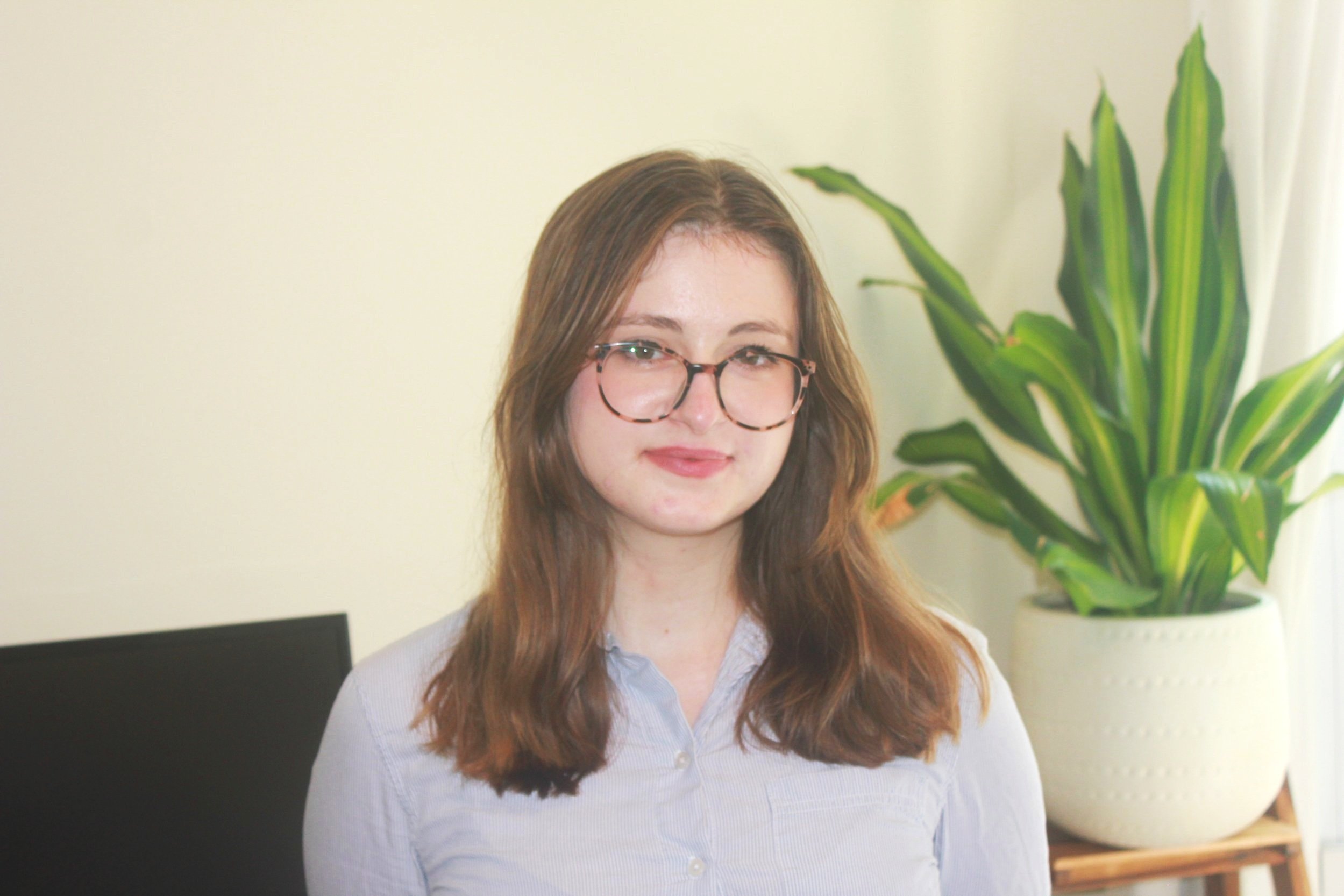 Headshot of me, brown hair, glasses, and a blue shirt standing in a room with a large potted plant.