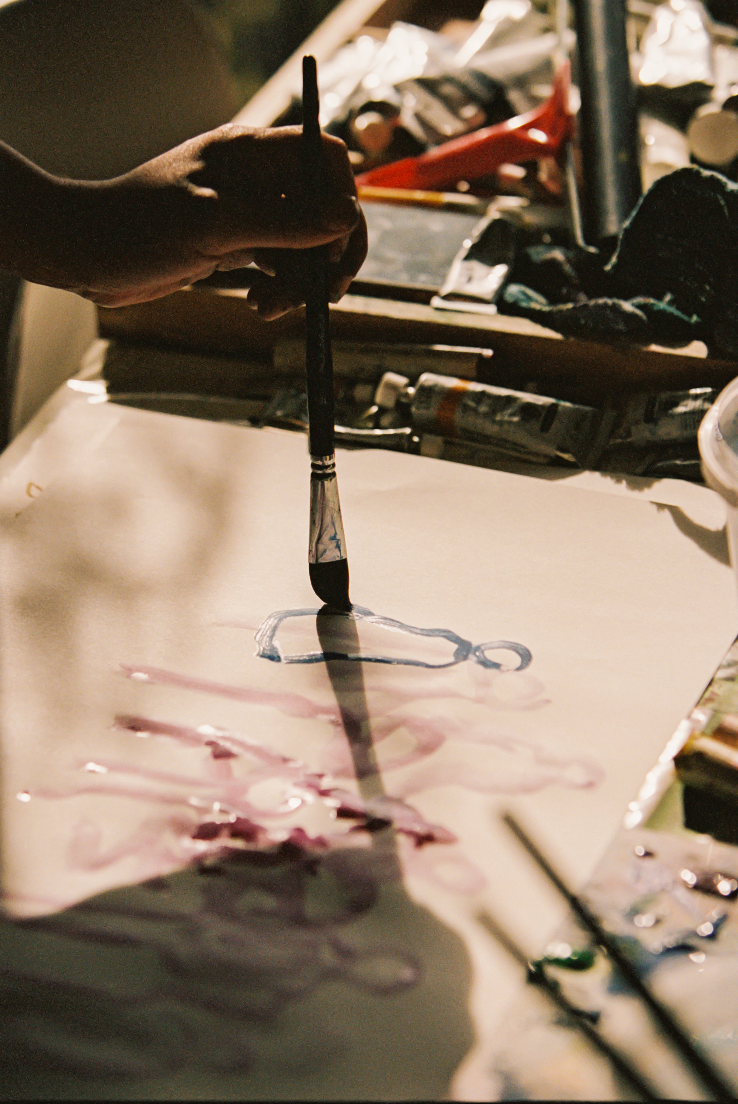 Person's hand using a paintbrush on a white canvas with colorful paints and art supplies in the background.