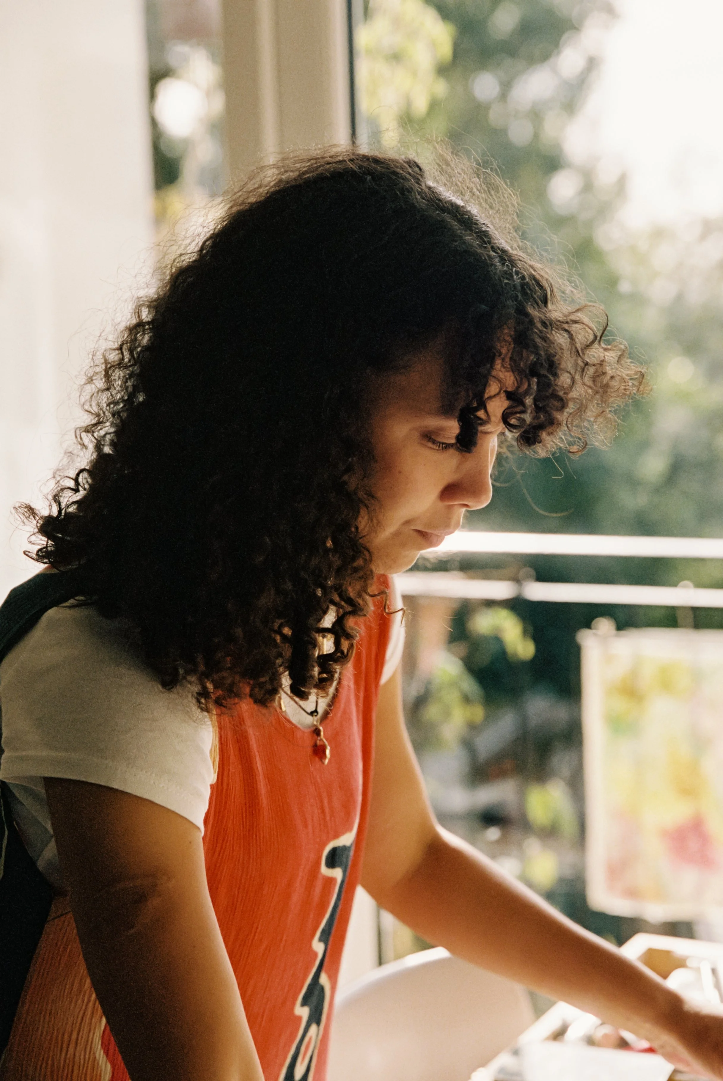 A person with curly hair wearing an orange tank top over a white shirt, looking down while standing indoors with a blurred background of a window and trees.