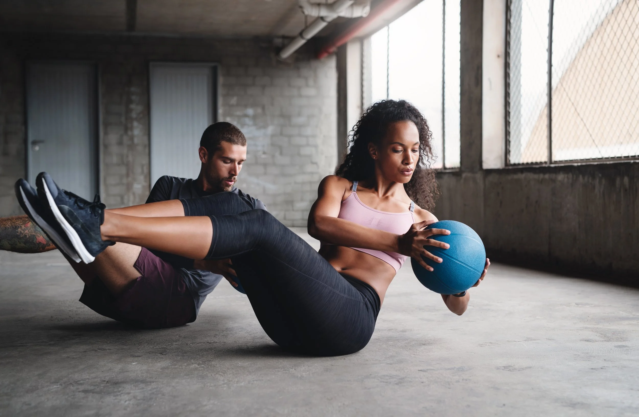 A woman in workout clothes performs a core exercise with a blue exercise ball while a man supports her legs in the healthstar club gym.