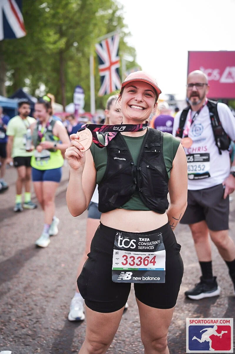 A woman smiles broadly after completing the London Marathon with medal in hand and british flags in the background