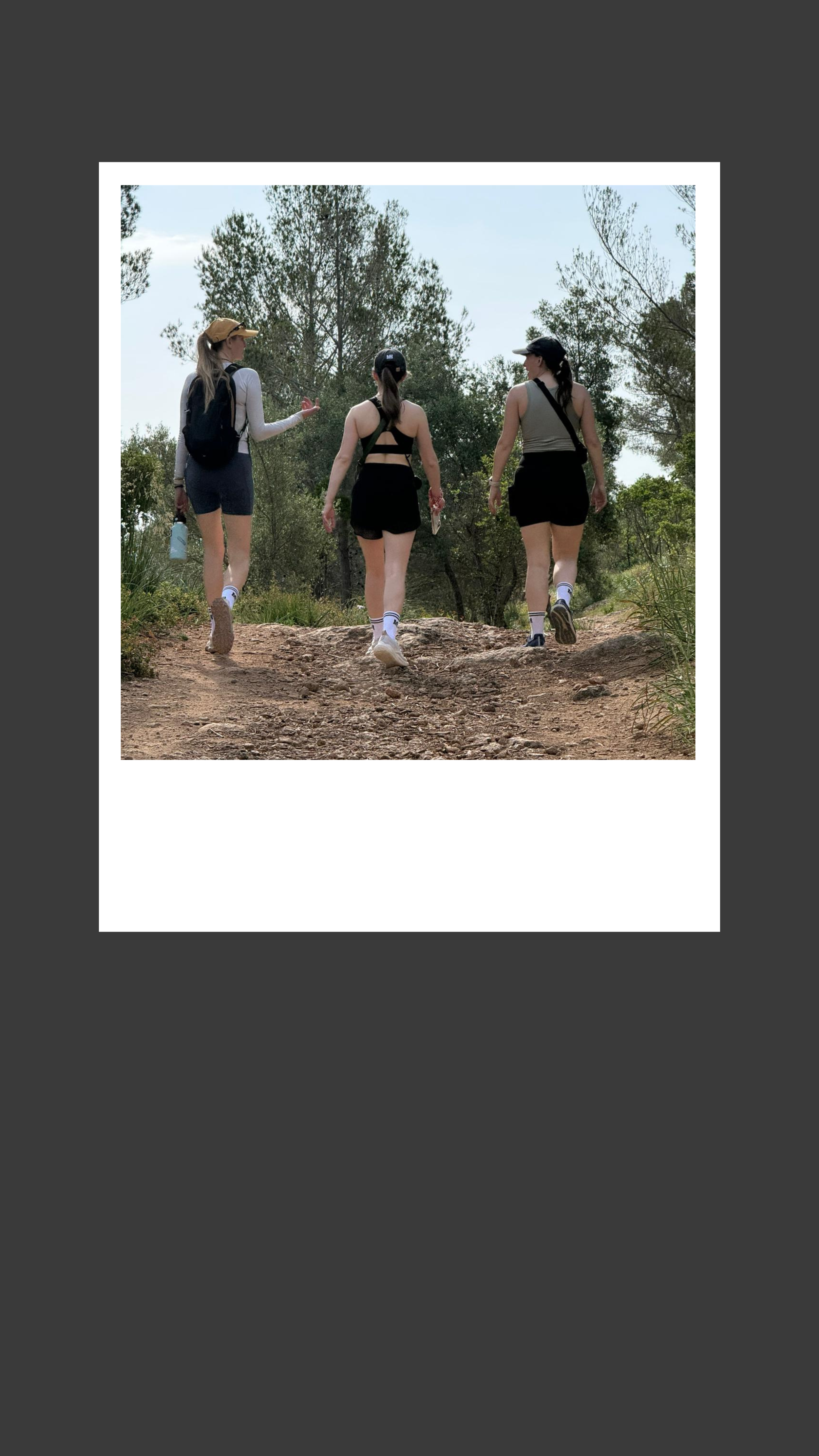 Three women hike on a dirt trail surrounded by trees in a forest, with one of them wearing a yellow hat and another carrying a water bottle.