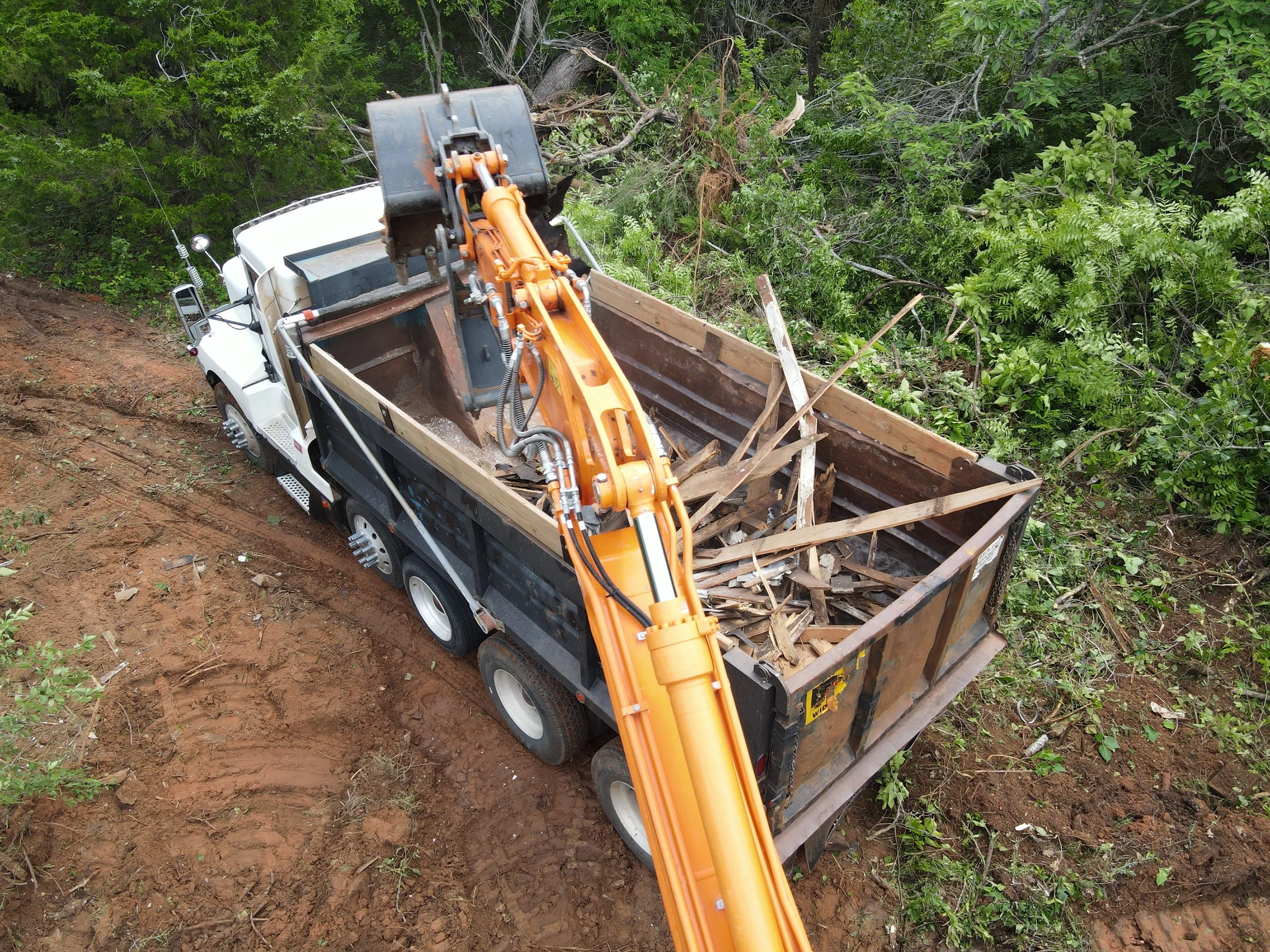 A truck with a boom attachment clearing trees and vegetation from a forested area, with debris and cut branches visible in the truck bed. Debris from a demolition
