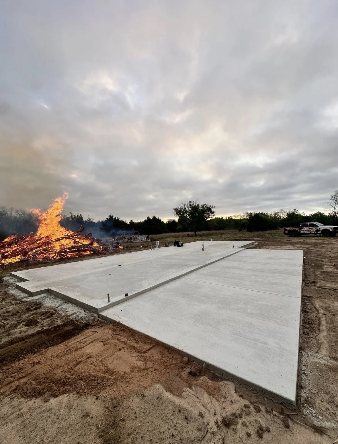 A newly poured concrete foundation slab on a construction site, with a burning fire and smoke in the background, along with a pickup truck and some tools.
