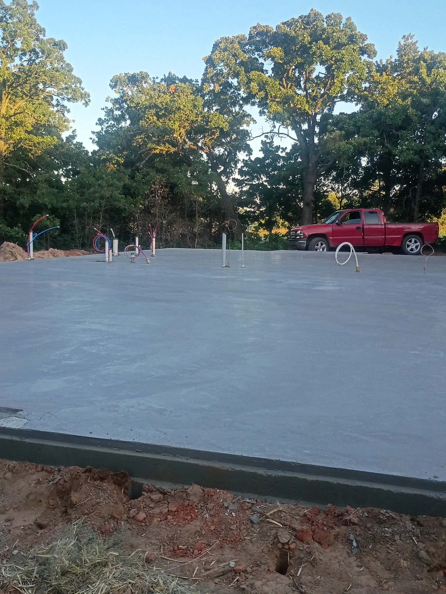 A newly poured concrete slab on a construction site with copper pipes protruding, a red pickup truck parked in a wooded area with large trees in the background.