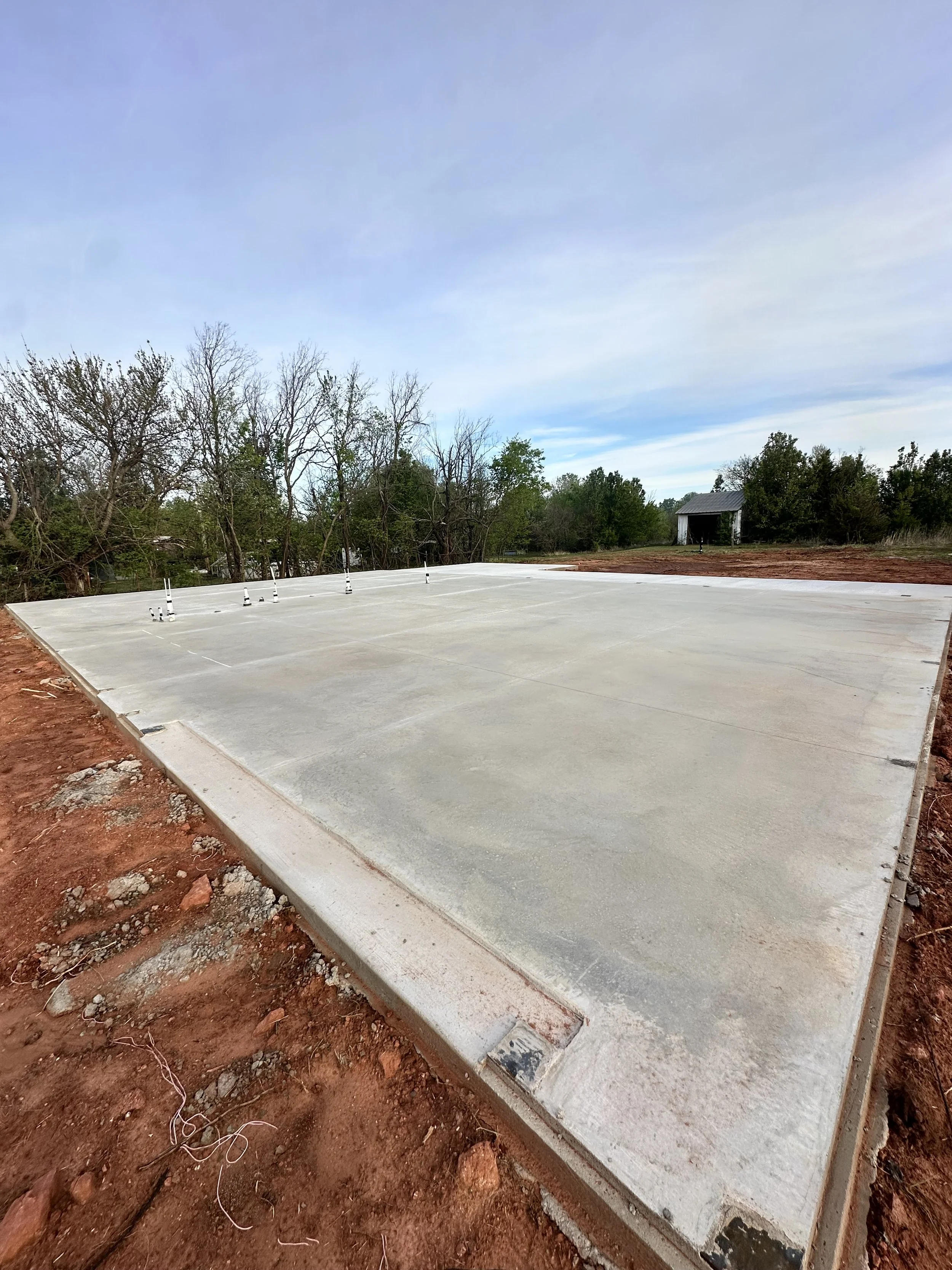A freshly poured concrete slab on a construction site, surrounded by red soil and some building materials, with trees and a small shed in the background.