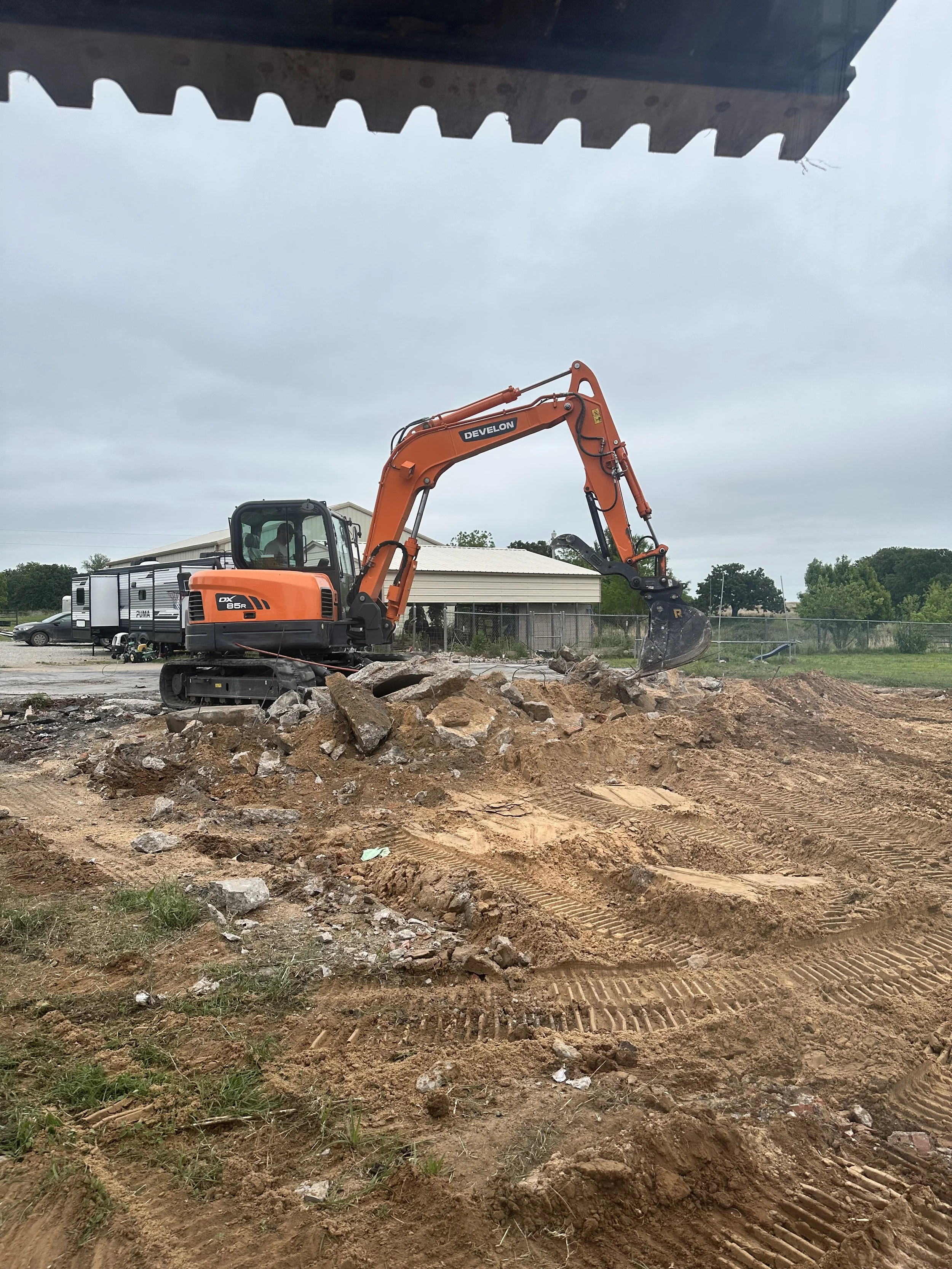 Construction site with an orange excavator moving dirt and rocks, tire tracks in the dirt, and a cloudy sky overhead.