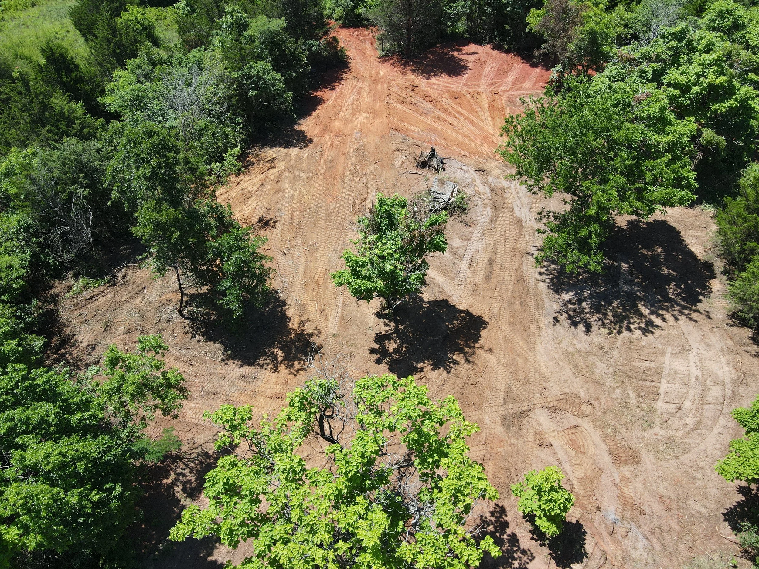 Aerial view of a cleared land area with reddish soil, surrounded by green trees and vegetation.