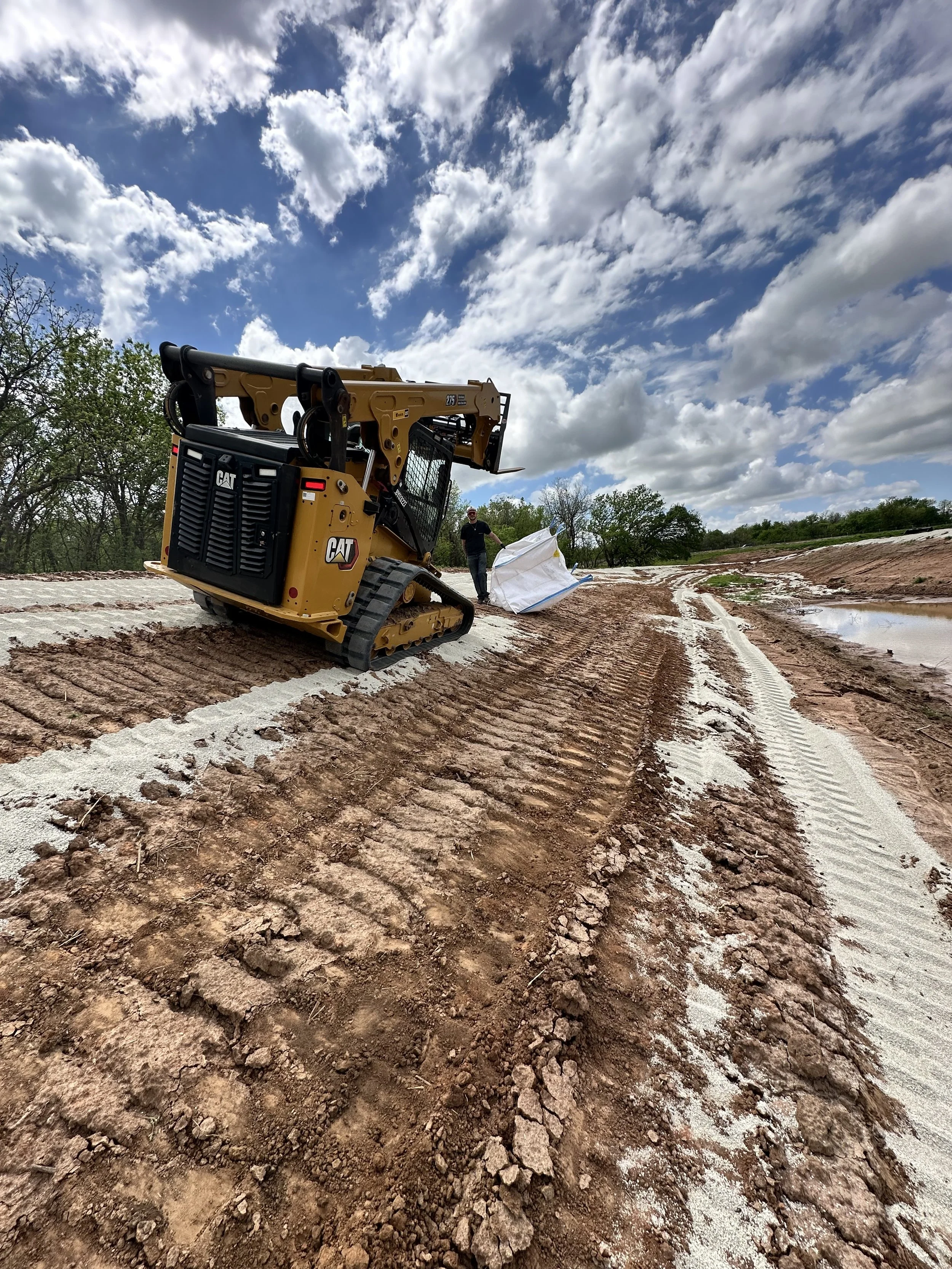A construction site with a large yellow Caterpillar skid-steer loader operating on a dirt terrain. A person alongside the equipment is holding a large white bag, with trees and a partly cloudy sky in the background.