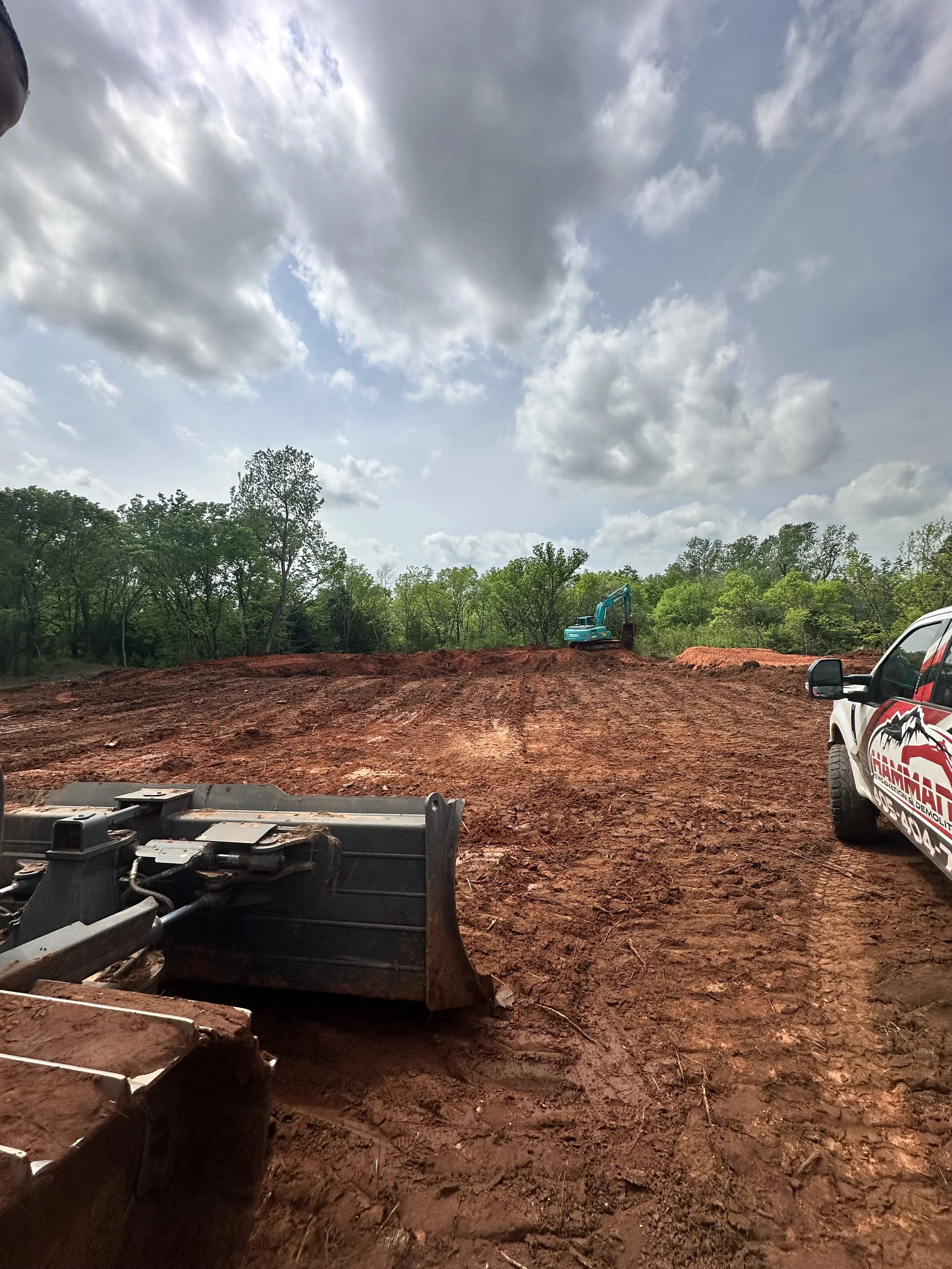 Construction site with cleared reddish dirt ground, a small blue excavator, and surrounded by green trees under partly cloudy sky.