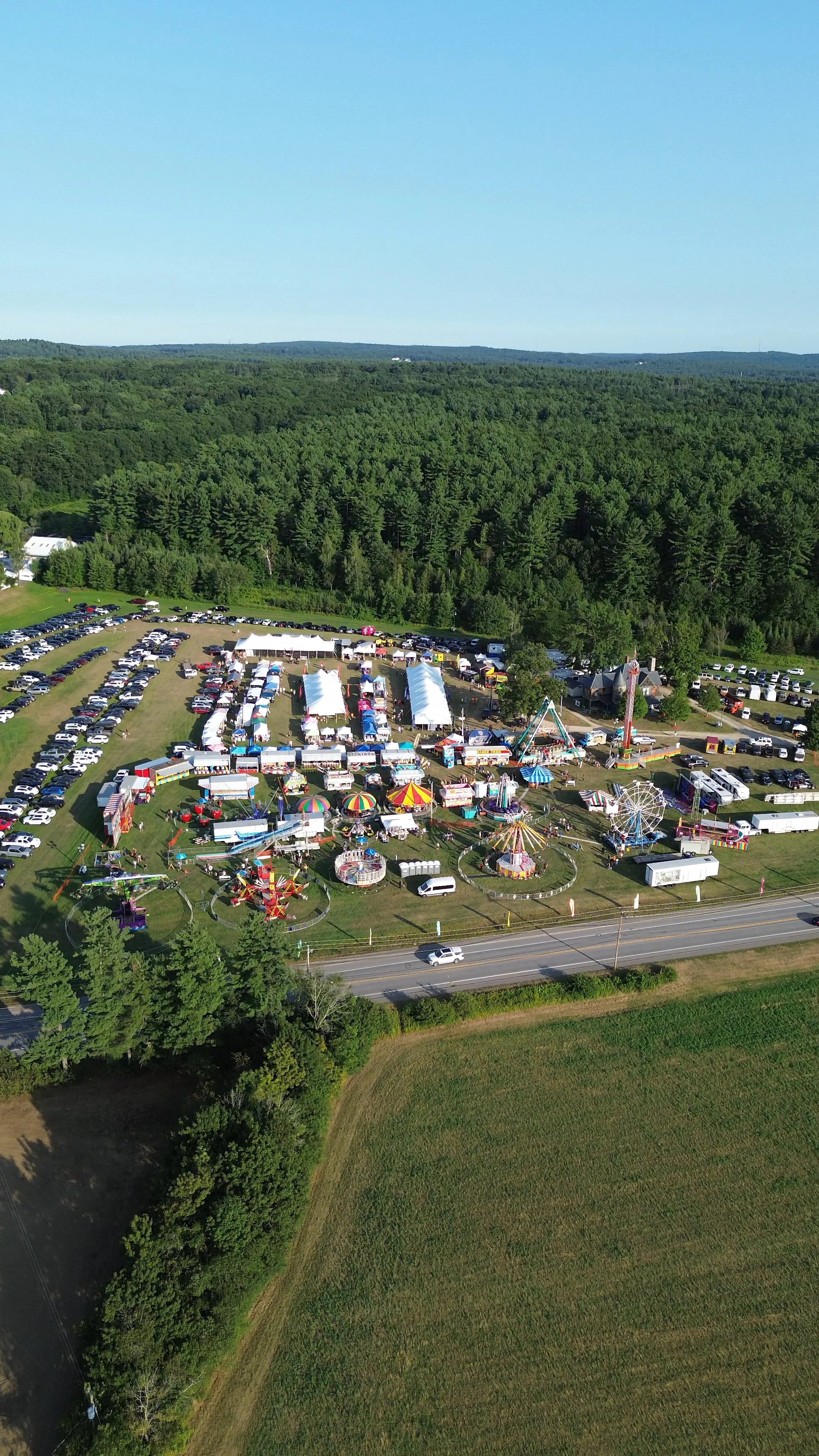 Aerial view of a carnival with rides, tents, and fairground attractions surrounded by trees and open fields, with some parked cars nearby.