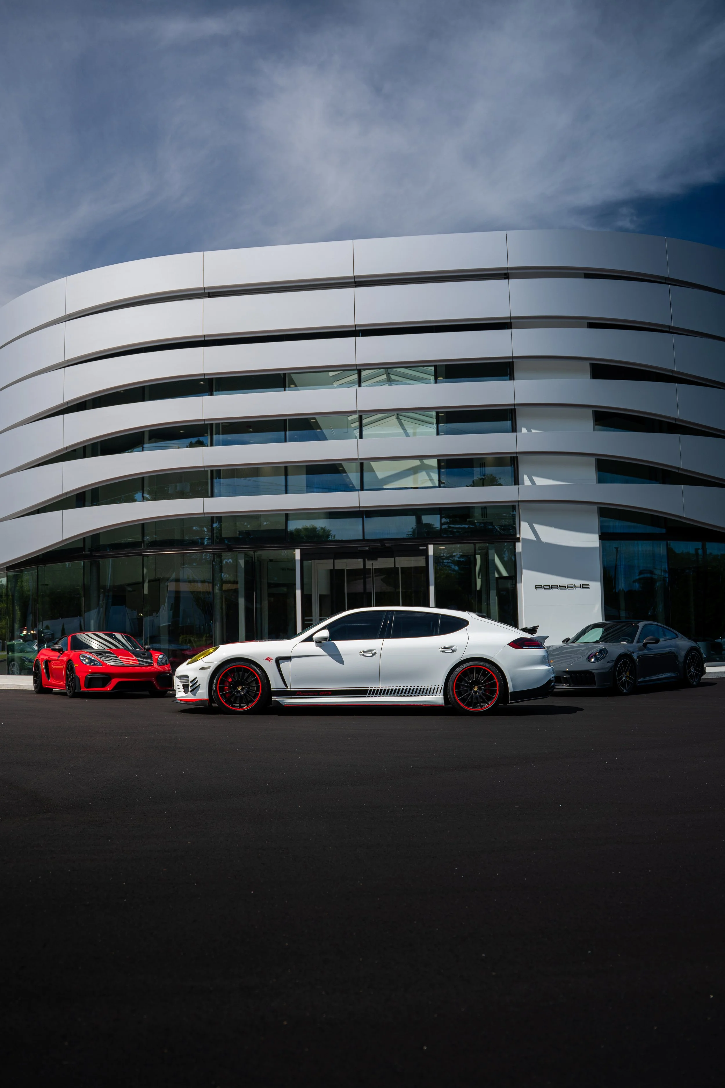 Three sports cars parked in front of a modern Porsche dealership building.