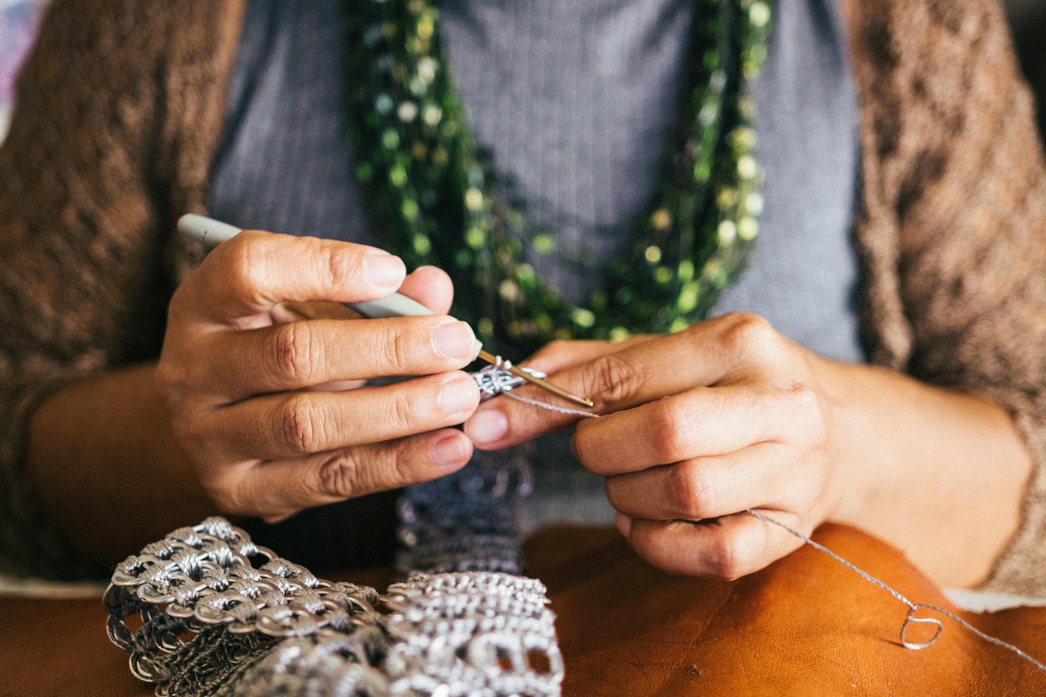Close-up of a woman's hands working on jewelry with a needle and thread, with jewelry pieces and beads on a wooden surface nearby.