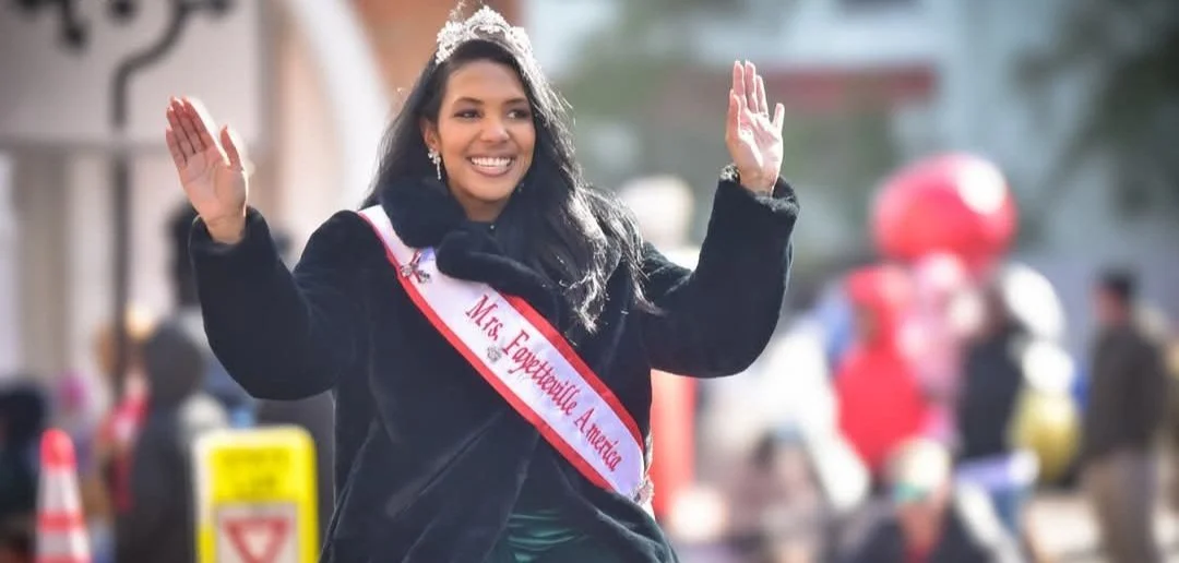 A woman wearing a sash and tiara, smiling and waving during a parade or celebration on a street, with blurred spectators and balloons in the background.