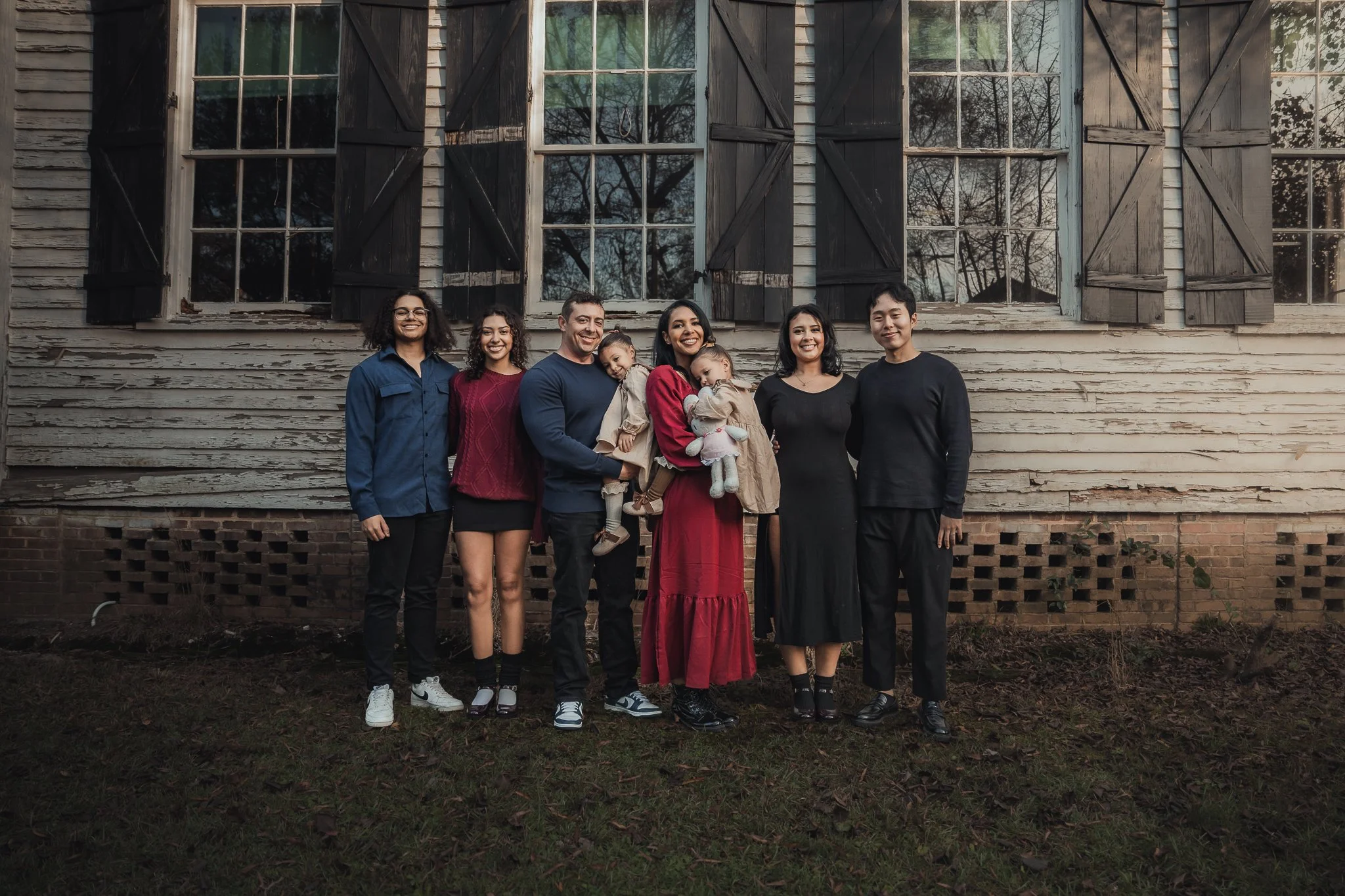 A diverse group of seven people, including children, standing together outside in front of an old, weathered house with peeling paint and dark shuttered windows, during daytime.