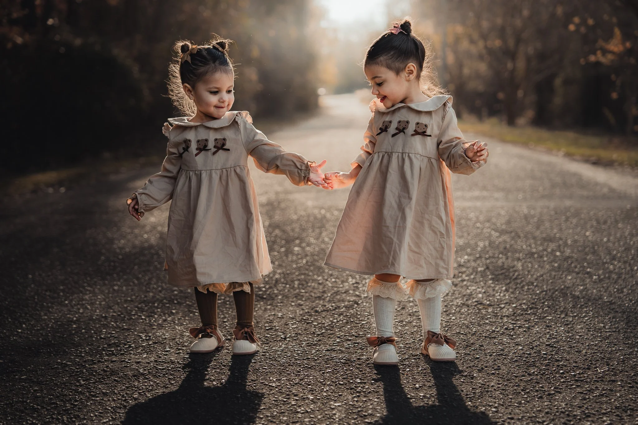 Two young girls wearing matching beige dresses with embroidered teddy bears, holding hands and smiling while standing on an outdoor road at sunset.