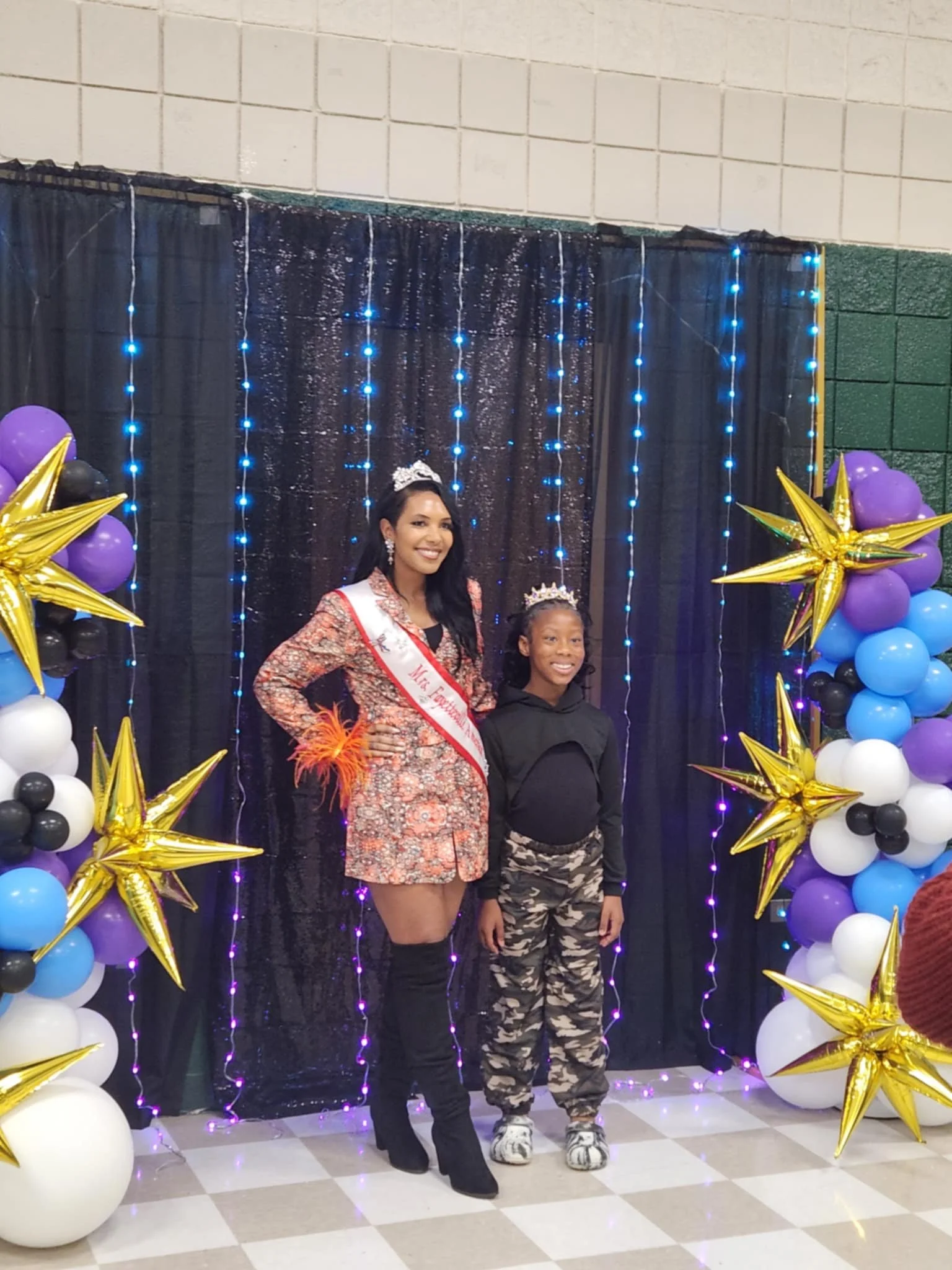 A woman and a girl standing together, smiling, at a celebration with balloon decorations and a black backdrop with blue string lights. The woman is wearing a sash, crown, and a floral dress with thigh-high boots. The girl is wearing camouflage pants,