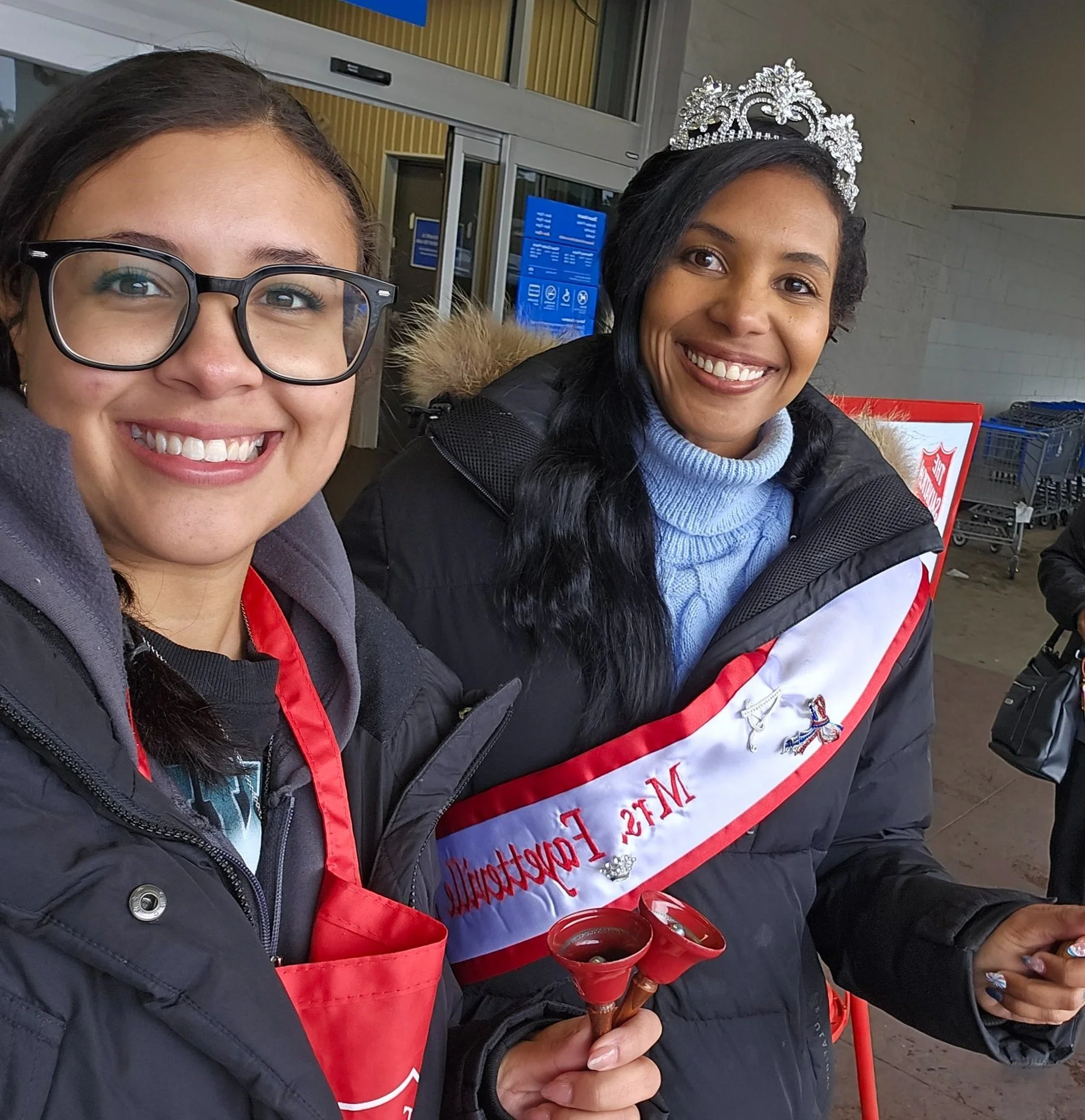 Two women smiling at the camera outside a building. One woman is wearing glasses, a red apron, and a black jacket, holding a small bell. The other woman is wearing a tiara, a sash that says 'Ms. Falcons,' a blue sweater, and a black jacket, also hold