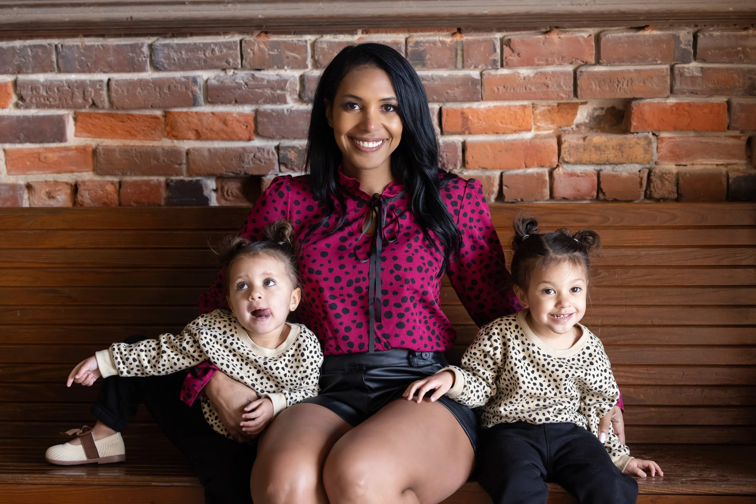 A woman sitting on a wooden bench with two young girls, all smiling, with a brick wall in the background.