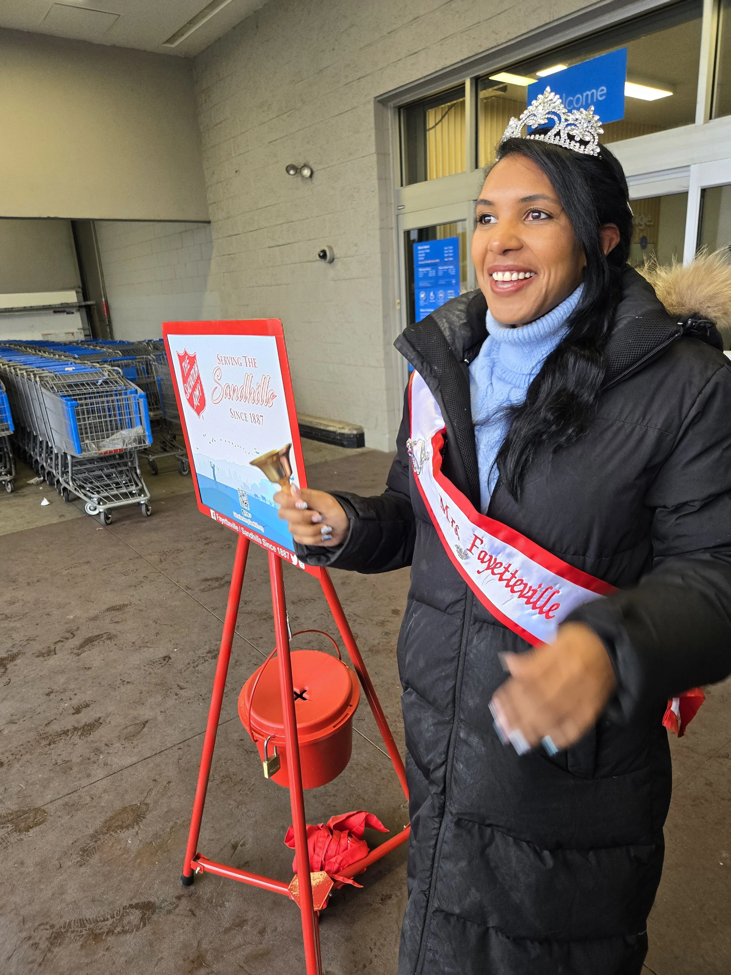 A woman wearing a tiara and a sash that reads 'Miss Fayetteville' is standing near a red bell and a sign that says 'Serving the Sandhills since 1887'. She is smiling and holding a small hammer, possibly about to ring the bell outside a building.