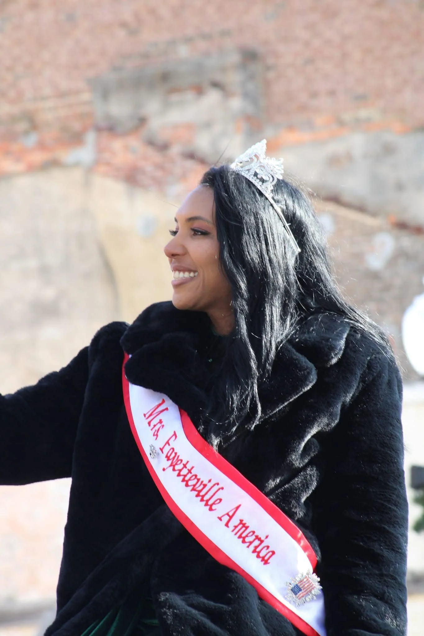 Woman with long black hair, wearing a tiara and black coat, smiling and wearing a sash that reads 'Miss Fayetteville America'.