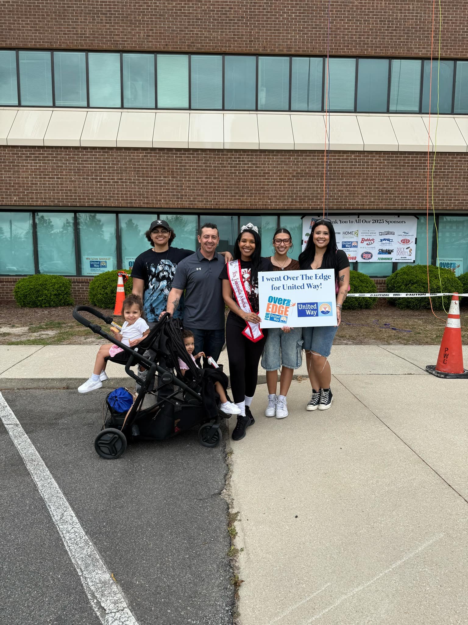 Group of five people, including three women, one man, and two young girls, standing outside a building, holding a sign that says, 'I went Over The Edge for United Way.' One woman is wearing a sash and tiara, indicating she may have participated in a 