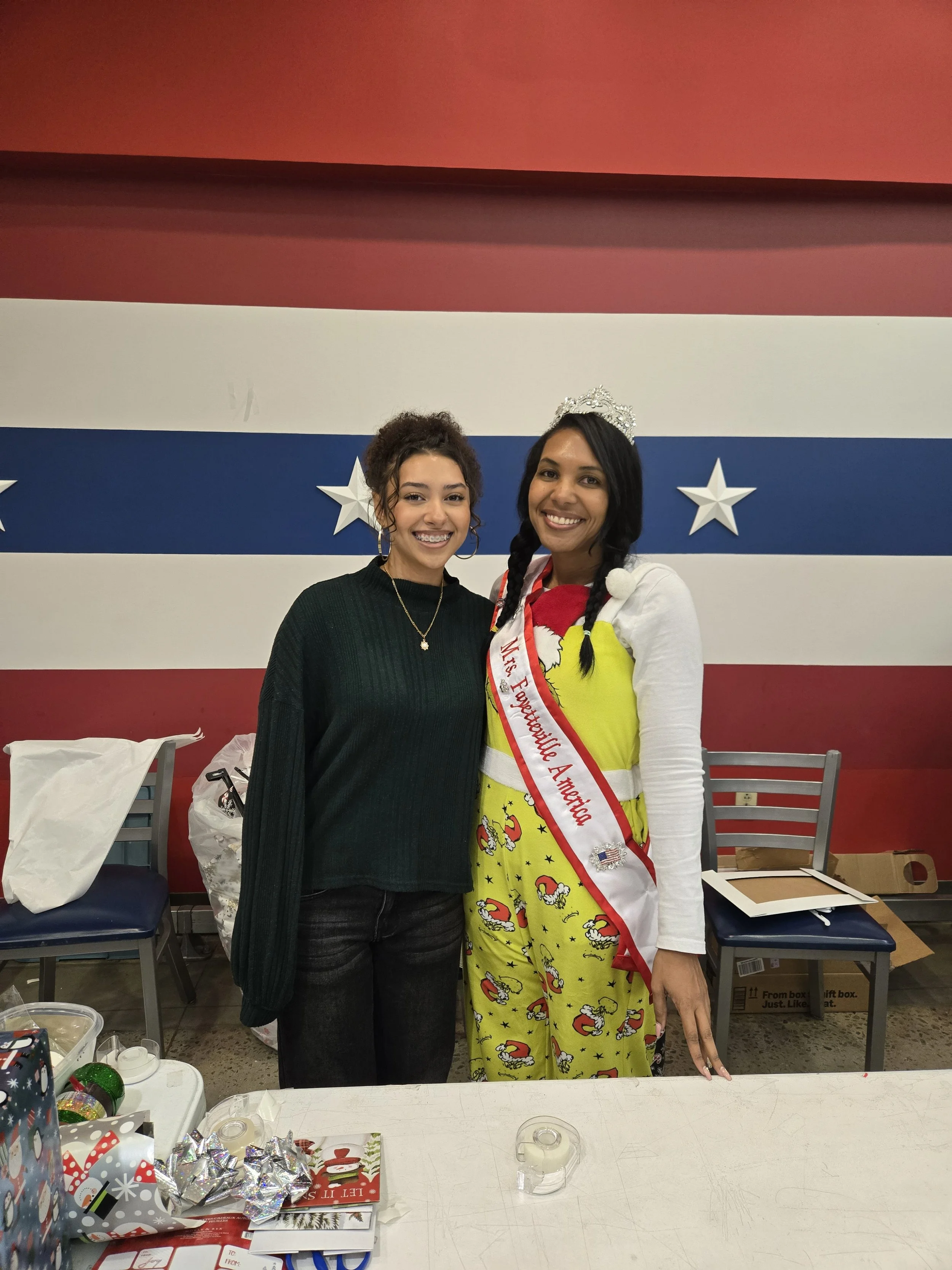 Two women smiling, one wearing a sash that reads 'Miss Fabulous America' and a tiara, standing in front of a red, white, and blue striped wall with stars, with gift-wrapped boxes and holiday decorations on the table in front of them.