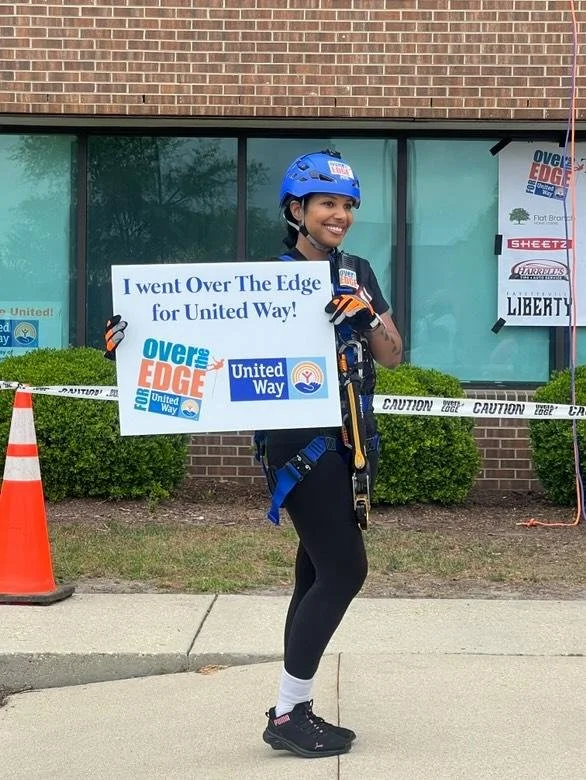 A young woman standing outside a building, wearing a safety helmet and harness, holding a sign that promotes unity and support for the United Way and Over The Edge event.