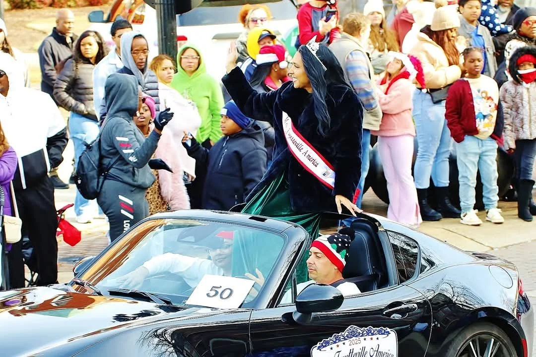 A woman wearing a sash that reads 'Beautiful America' is standing in a black convertible car, waving to a crowd during a parade. The car has a sign with the number 50 on it, and there is a man wearing a Christmas-themed hat sitting in the passenger s