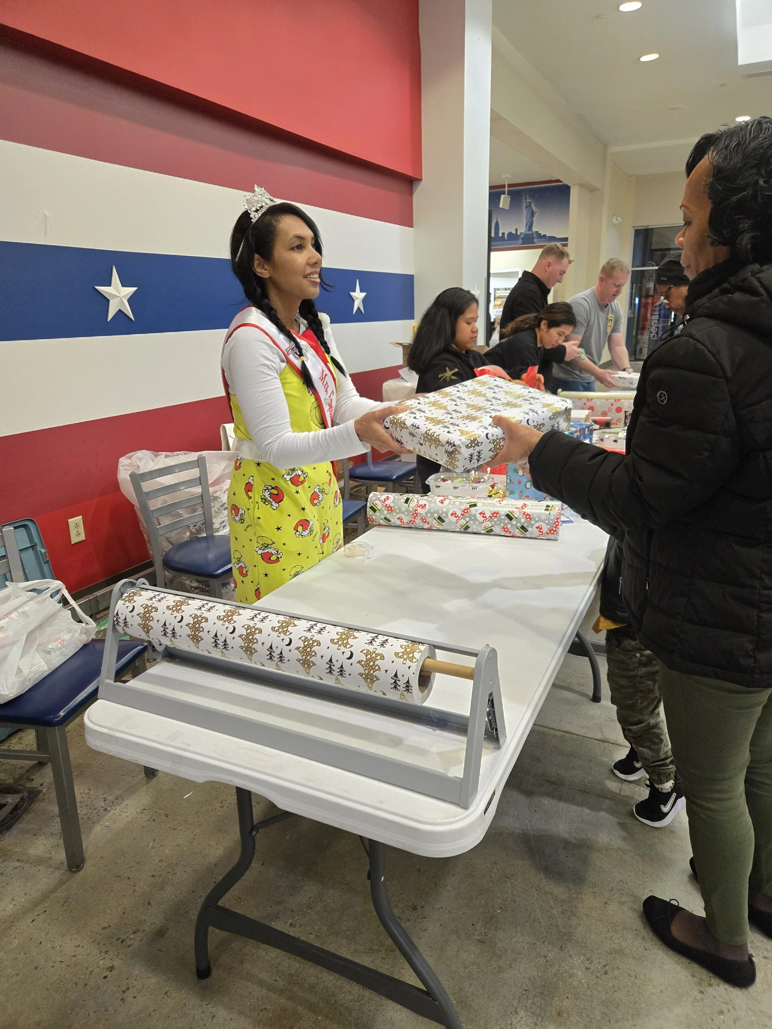 A woman wearing a Miss America sash and a tiara is handing a wrapped gift to a woman in a black jacket at a gift-wrapping station, with others in the background wrapping presents.