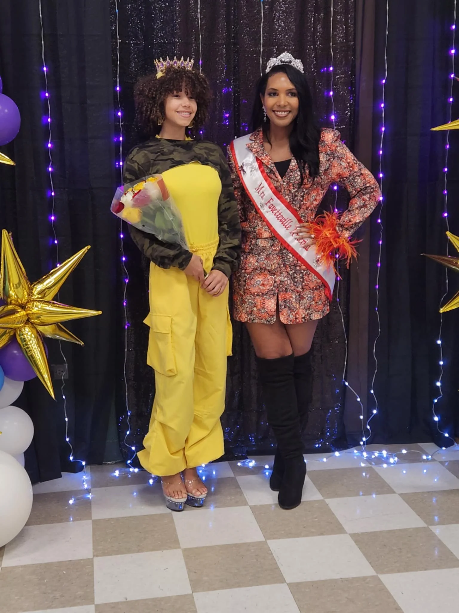Two women standing side by side, one wearing a crown and holding a bouquet of flowers, the other wearing a sash that reads 'Mrs. Fayetteville 2023,' and a tiara, at a celebration event with black and purple background drapes and gold star and balloon