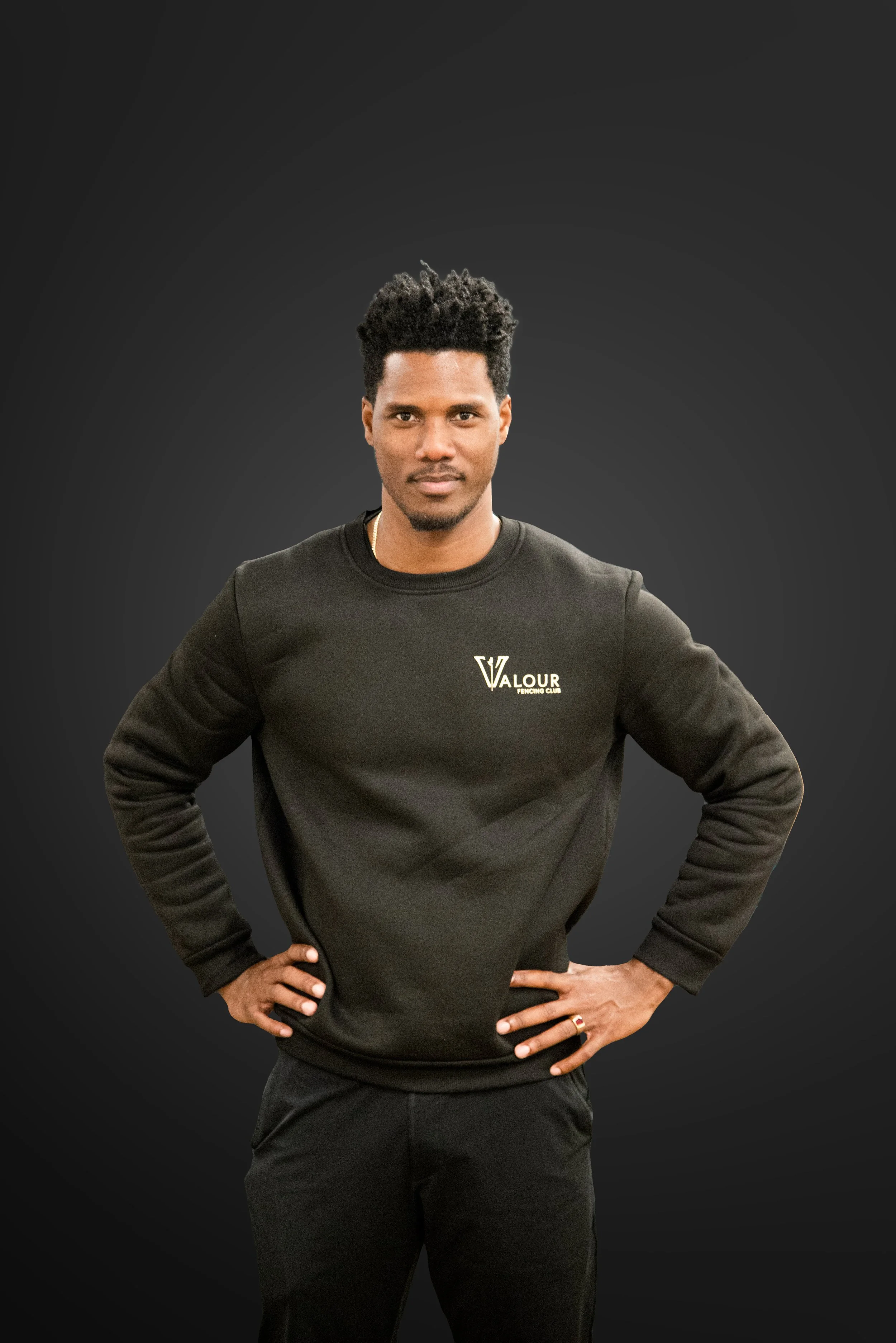 A man with dark hair and a goatee wearing a black sweatshirt with 'Valour Fencing Club' logo, standing with hands on hips against a dark gradient background.