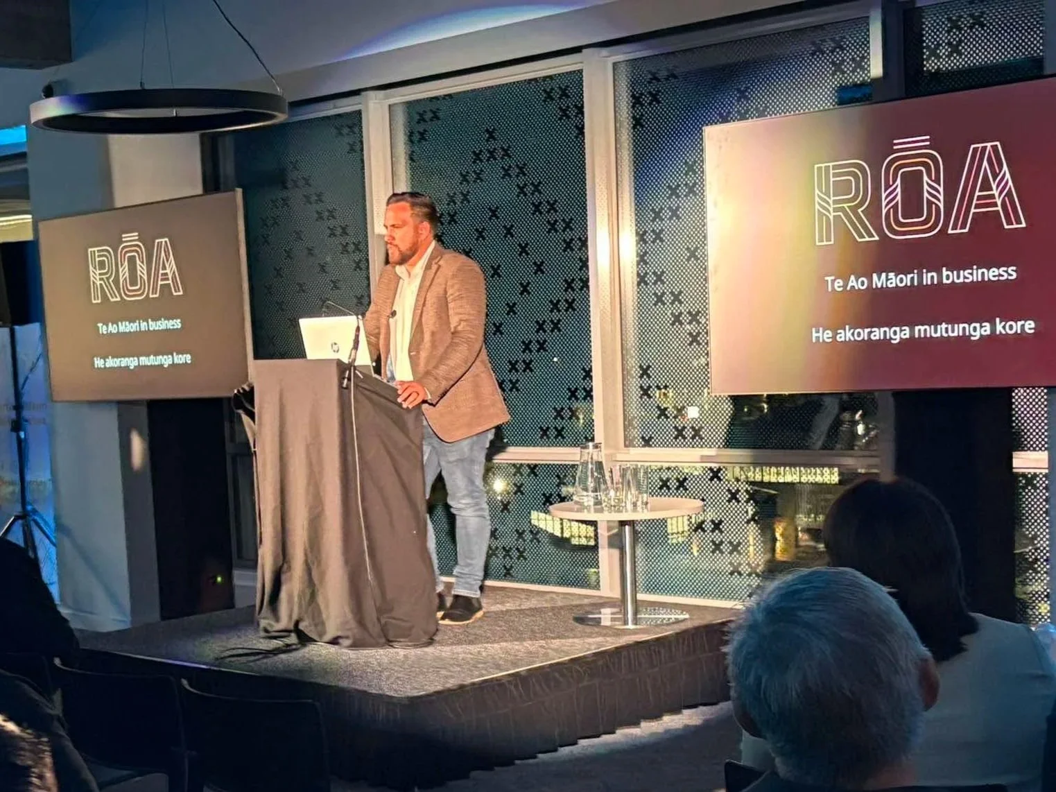 A man in a brown blazer and jeans giving a presentation on a stage with two large screens displaying the word 'ROA' and text in Maori. The audience is visible in the foreground.