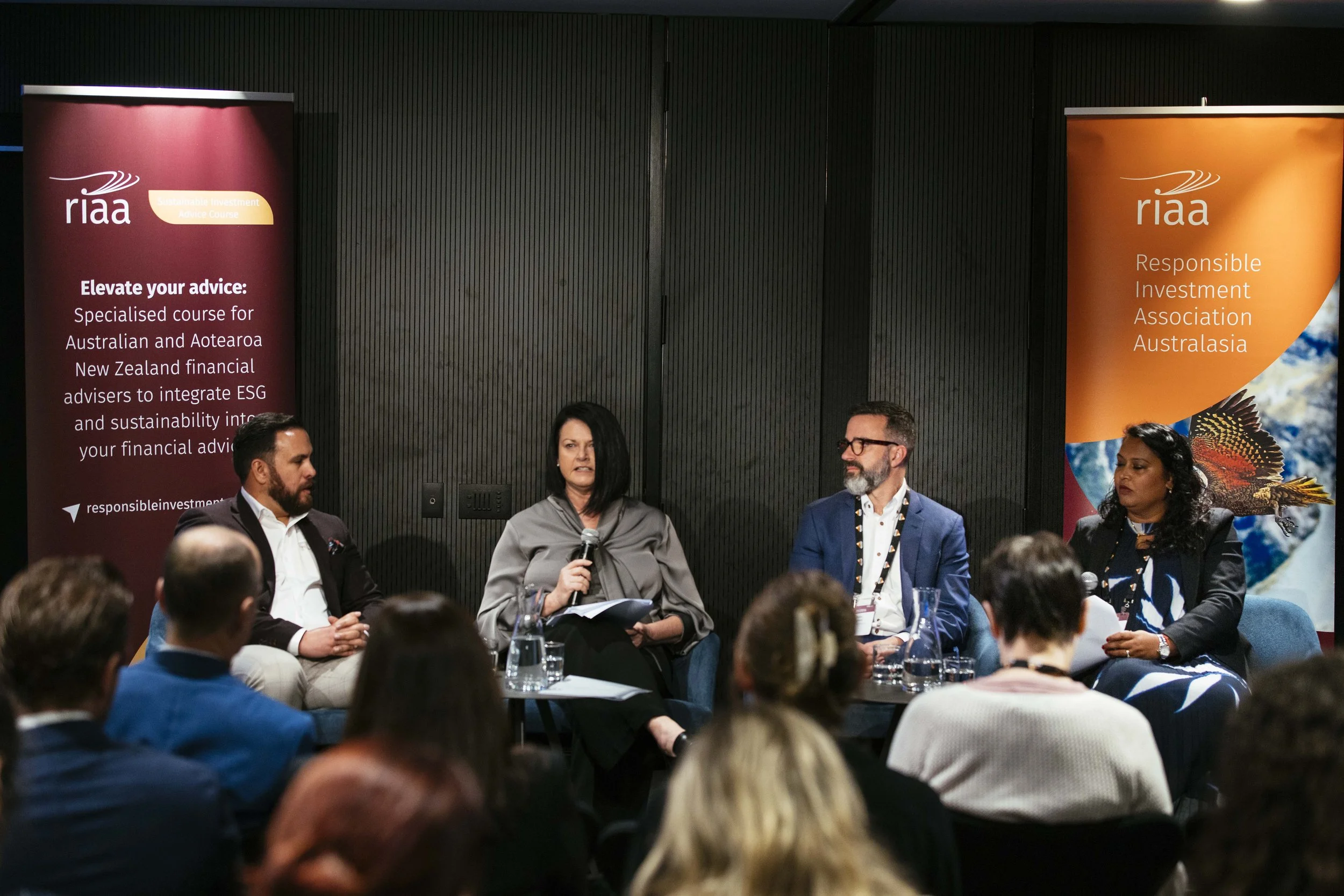 Panel discussion at a conference with four speakers seated on stage, two men and two women, with audience members listening. Two banners behind them display 'RIAA' and slogans about investment and sustainability.