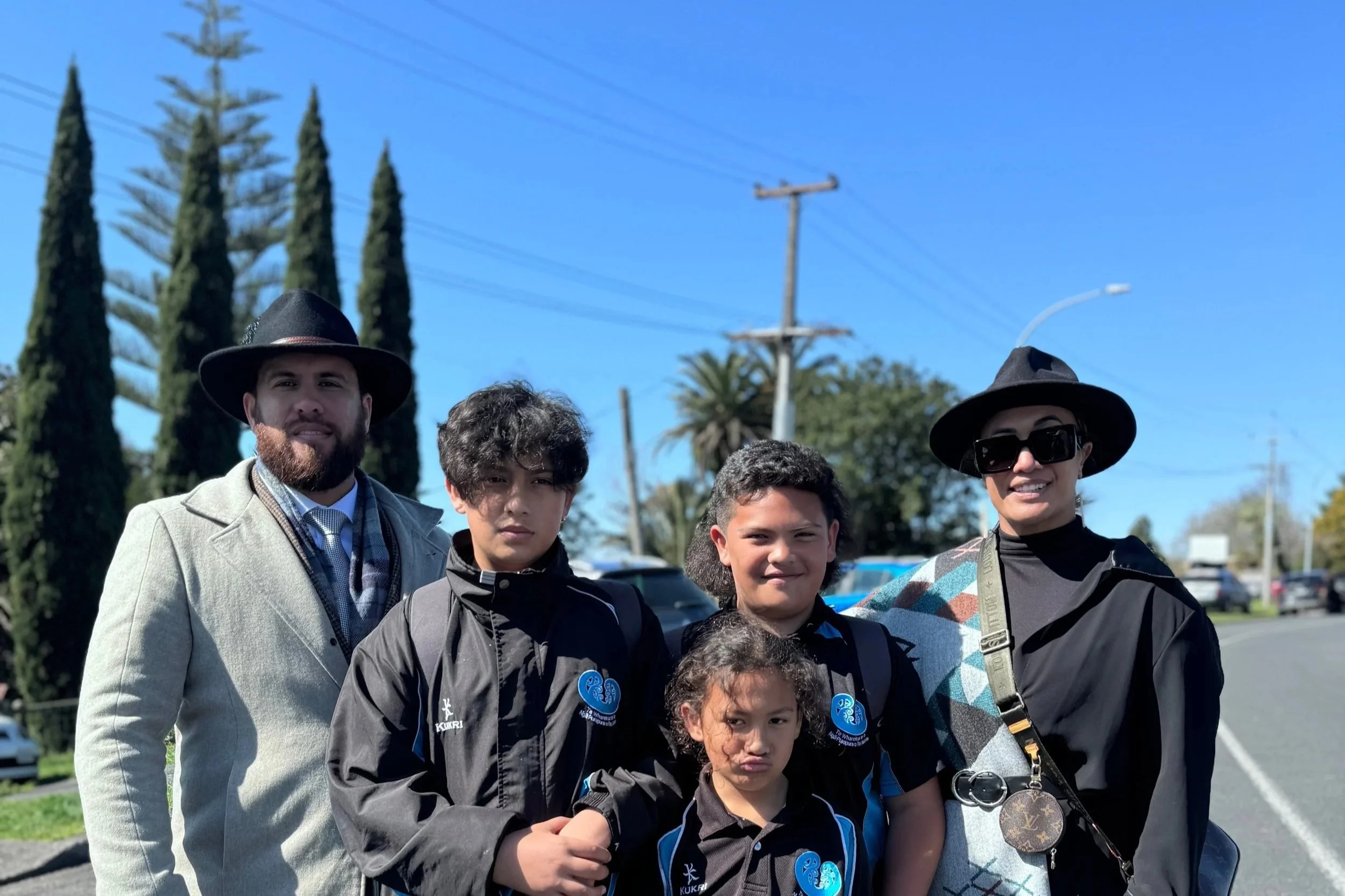 Group of six people, including children and adults, standing outdoors on a sunny day with trees and power lines in the background. The people are dressed in casual and semi-formal clothing, some wearing hats and sunglasses, smiling at the camera.