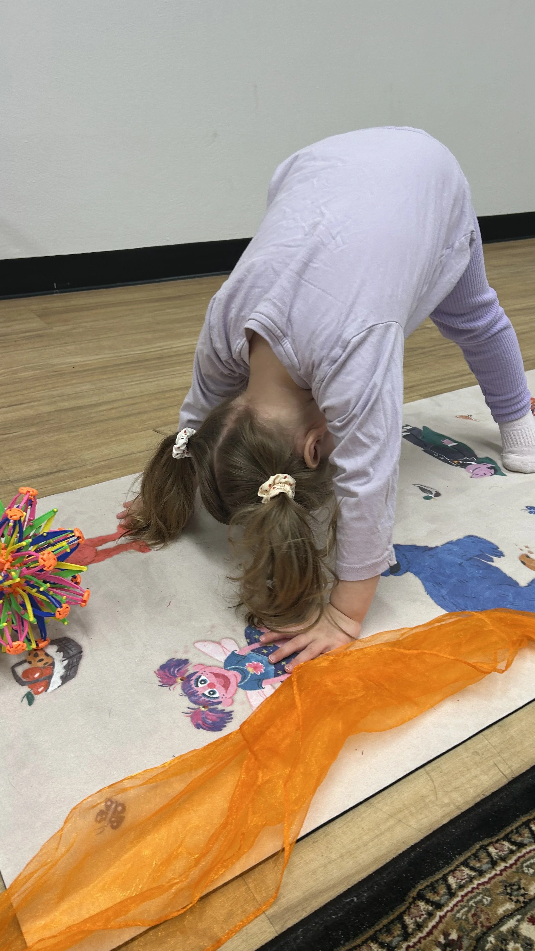 Young child in pink sweatshirt and striped leggings performing a forward fold in a grassy oudoor area.