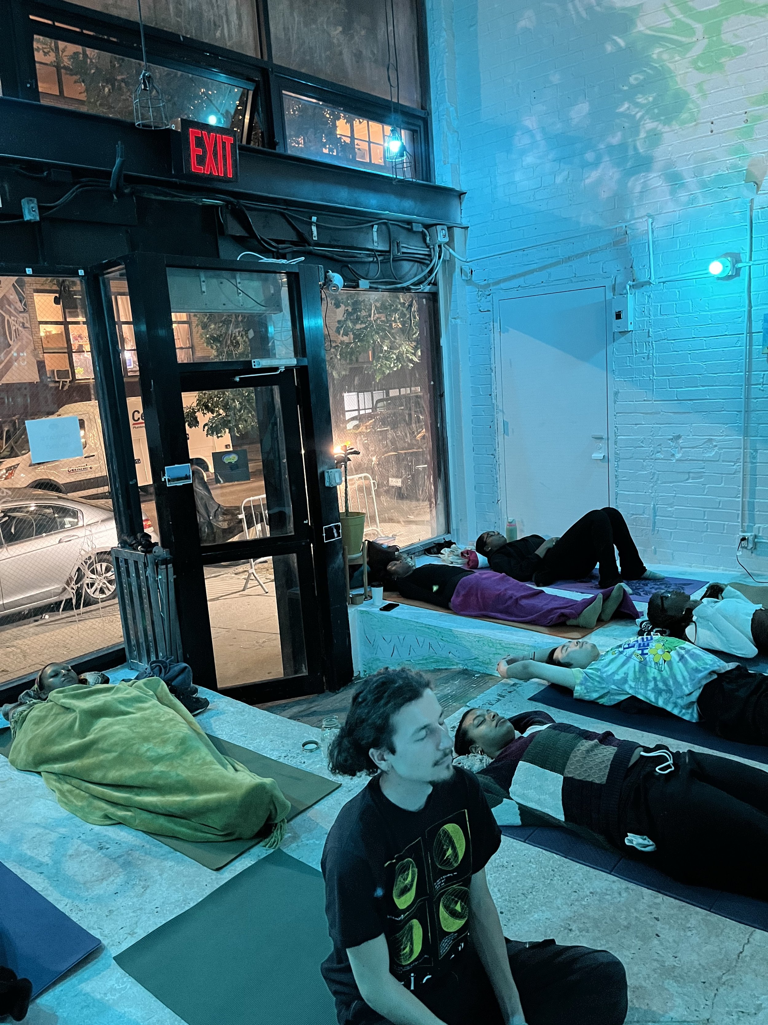 People practicing yoga or meditation lying on mats inside a brightly lit indoor space with a large window showing parked cars outside. A woman is seated cross-legged on a mat in the foreground